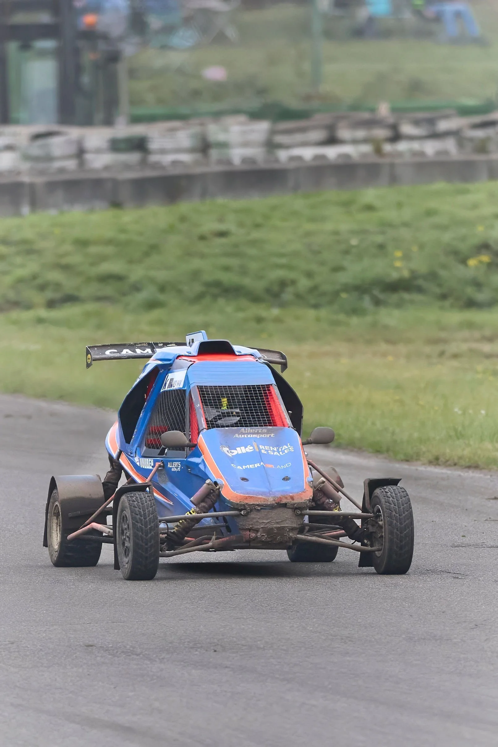 A blue and black race car with a red roll cage, driving on a paved track with grass in the background.