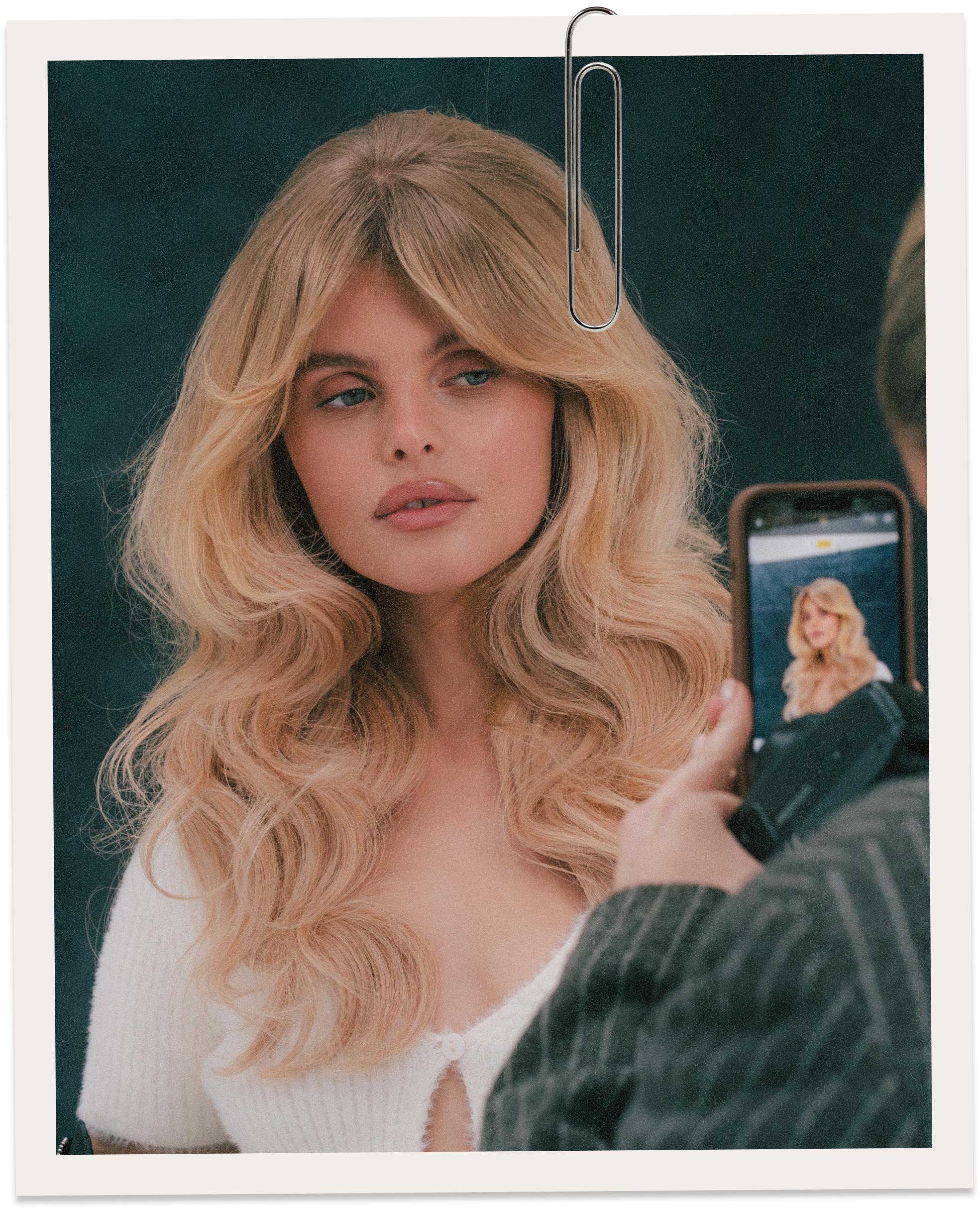 A woman with long, wavy blonde hair and light makeup poses for a photo taken by a person holding a smartphone, with a dark background.
