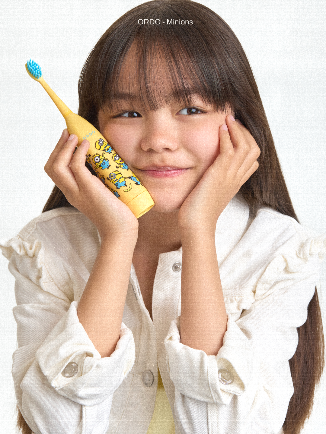 A young girl with long brown hair and a fringe, she is holding a yellow electric toothbrush with blue bristles and Minions graphics on the handle.