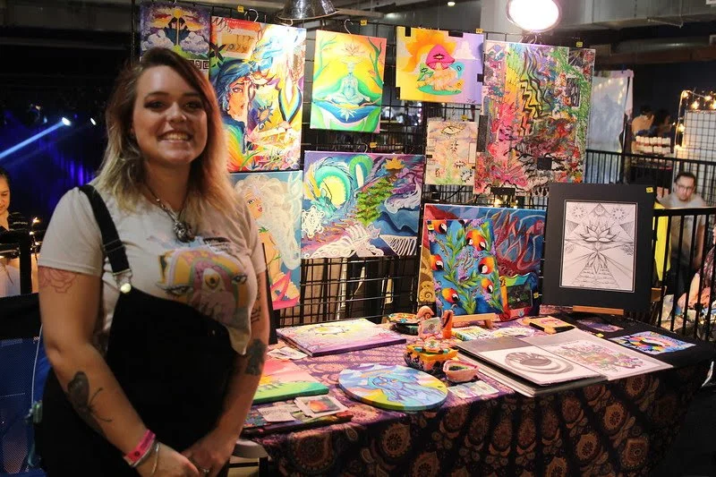 A woman with blonde hair and a white t-shirt featuring a colorful art design, standing in front of a display table with vibrant, colorful artwork and illustrations at an art fair or exhibition.