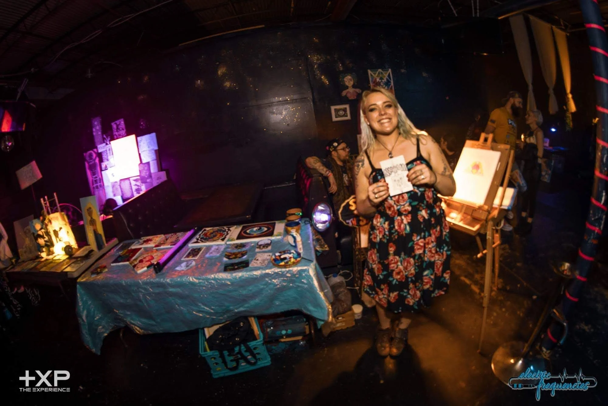 A woman with blonde hair, tattoos, and a floral dress smiling and holding artwork in a dimly lit art event or festival with paintings, artwork, and other attendees in the background.