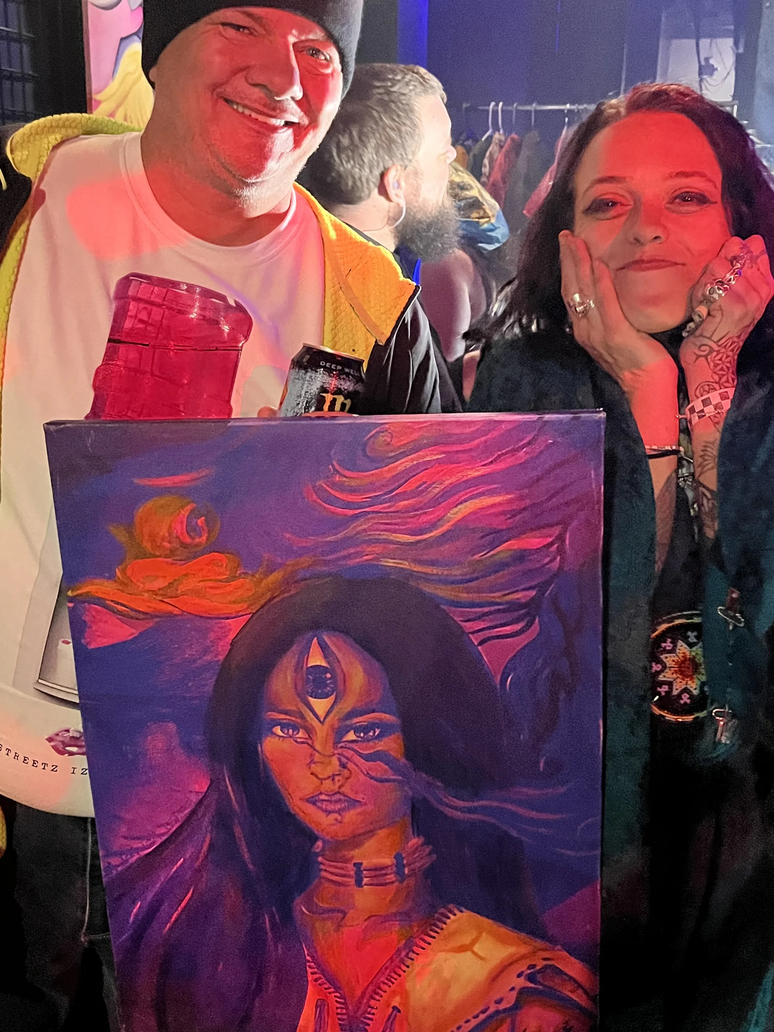 Two people smiling and posing for a photo at an event, holding a vibrant, surreal portrait painting of a woman with long dark hair, a third eye on her forehead, and vivid colors like purple, orange, and pink.
