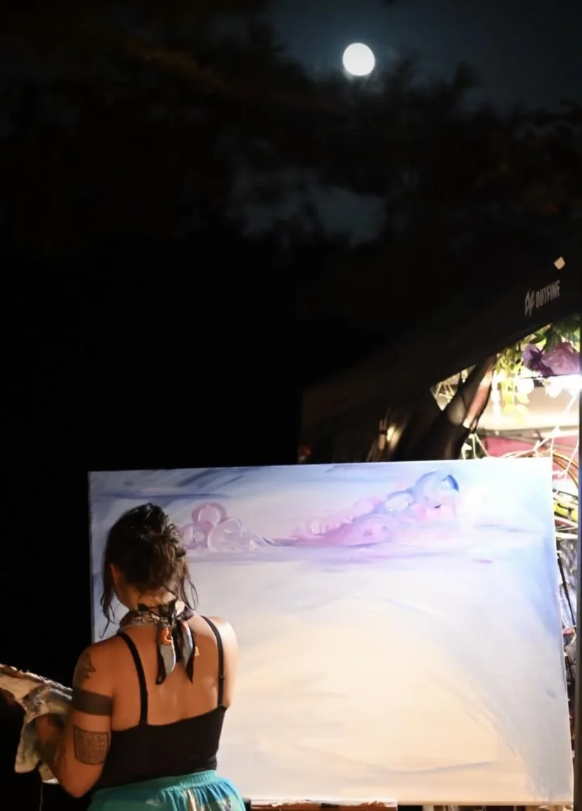 A female artist with tattoos, wearing a black tank top and a bandana around her neck, painting a landscape on a large canvas outdoors at night under the moon and trees.