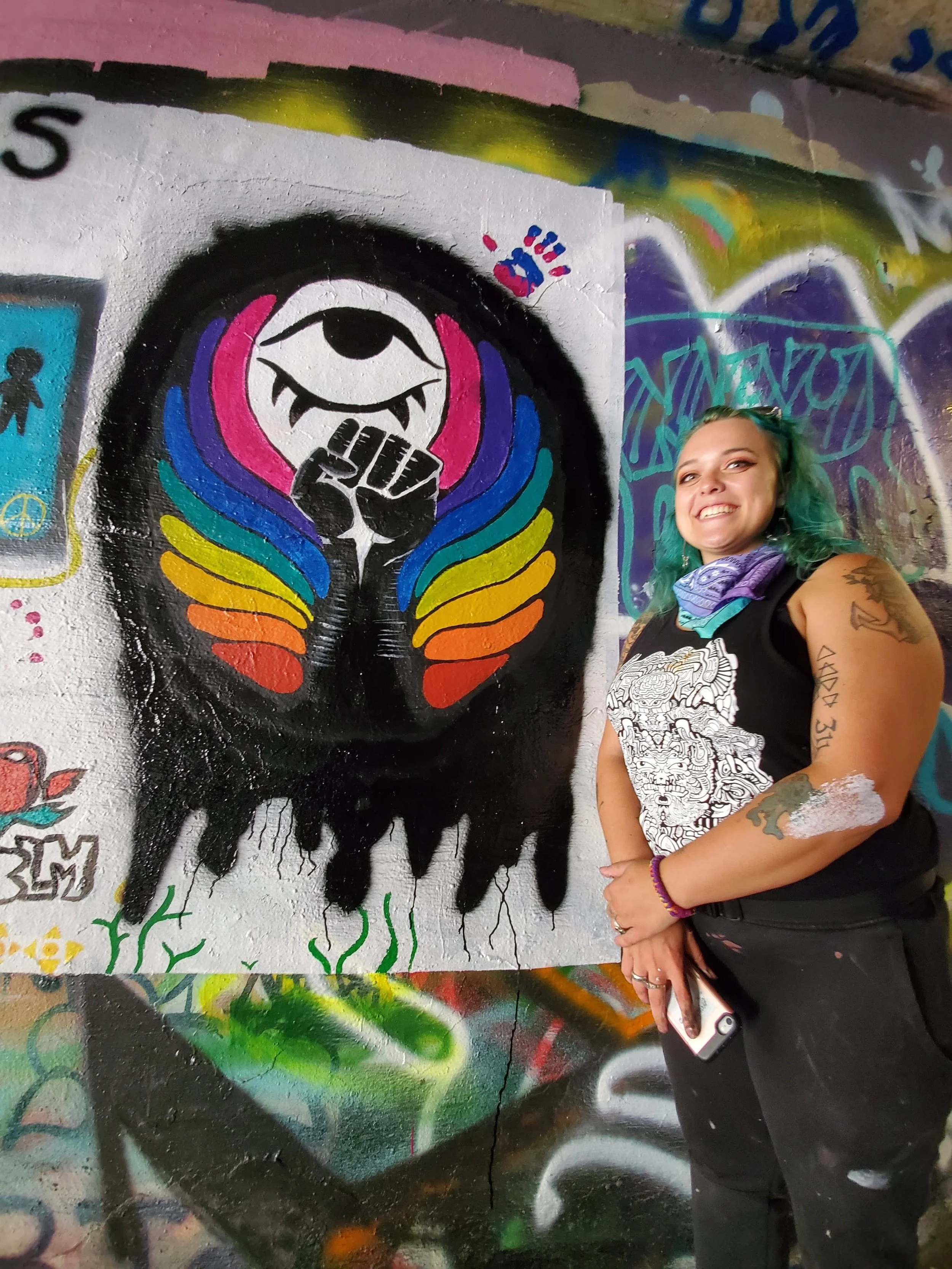 A young woman with green hair and tattoos smiling next to a colorful street art mural of an eye with a raised fist inside it, surrounded by rainbow-colored wings.