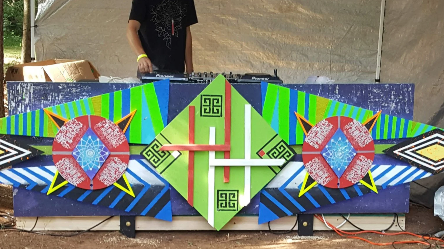 Colorful geometric artwork on a large board behind a DJ at an outdoor event, with a person wearing black with a yellow wristband.