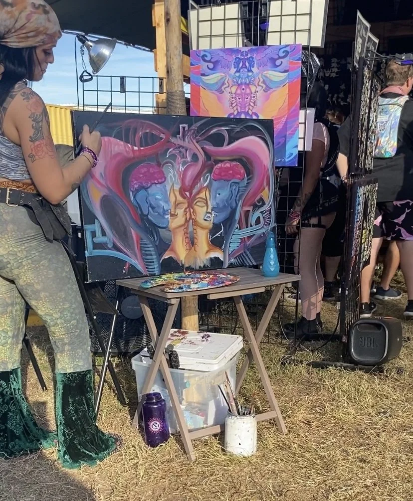 An artist at an outdoor art fair paints a colorful, surreal picture of two blue-skinned figures with pink brains connected to a central face with flaming pink hair. Other artworks and people browsing booths are visible in the background.