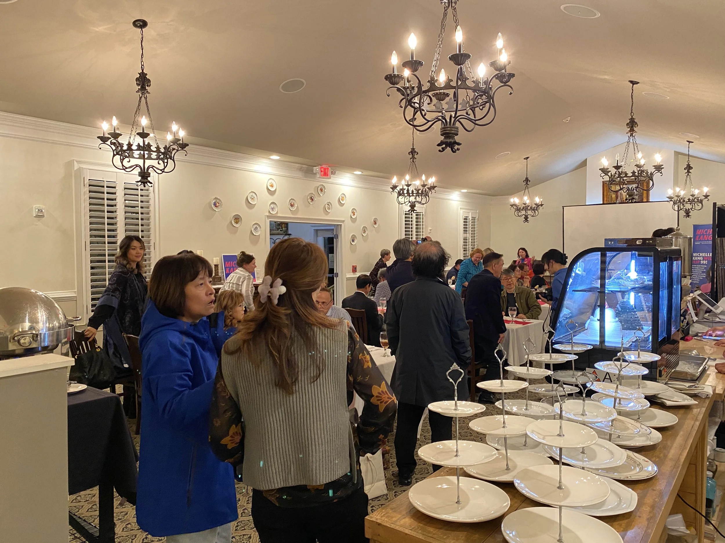 Crowd gathered at a social event in a banquet hall with chandeliers, white walls with decorative plates, and a table with stacked plates and a display case.