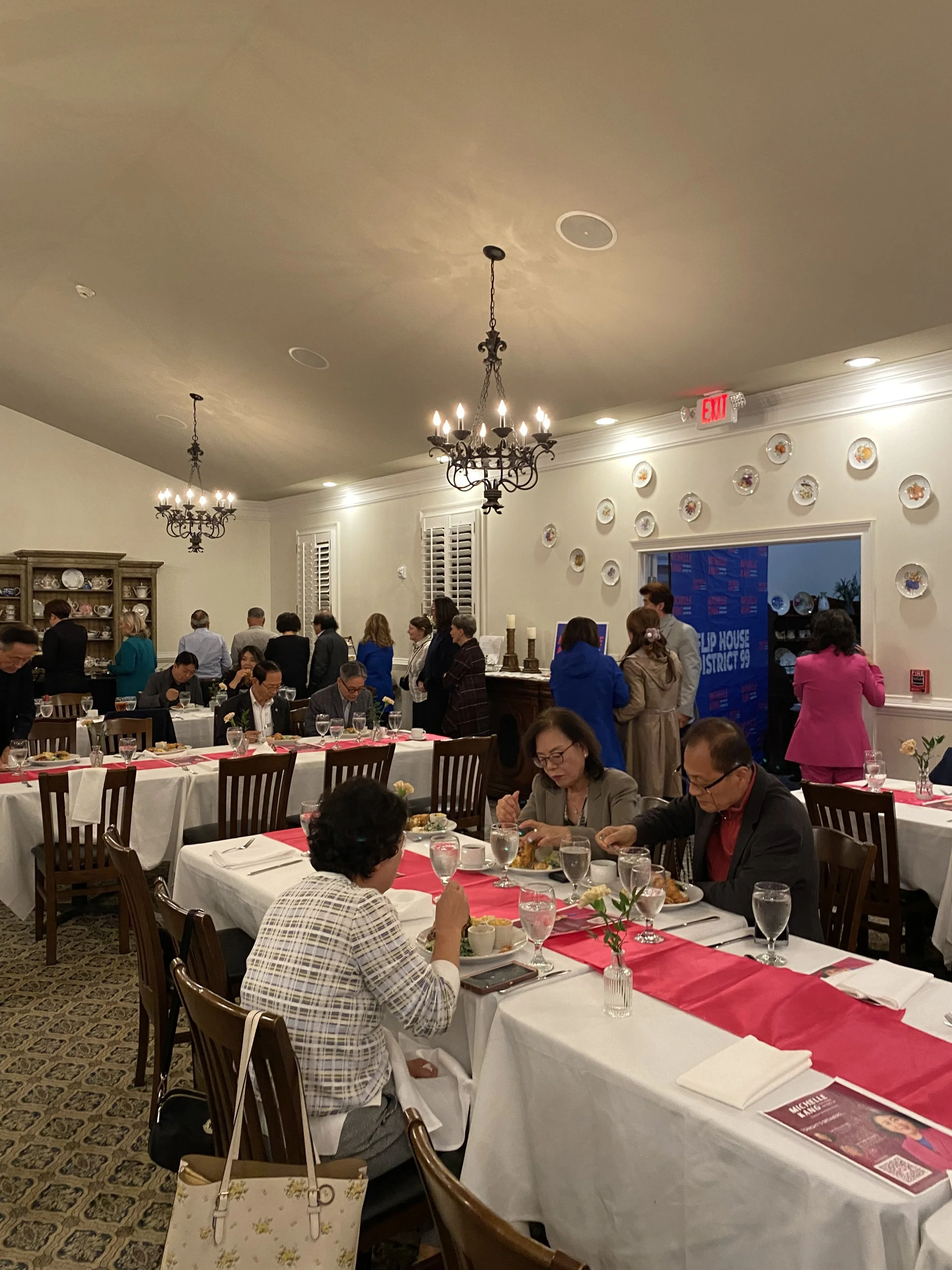 People dining and socializing in a formal dining room with chandeliers, tables with white tablecloths and red table runners, and walls decorated with plates and shelves of dishes.