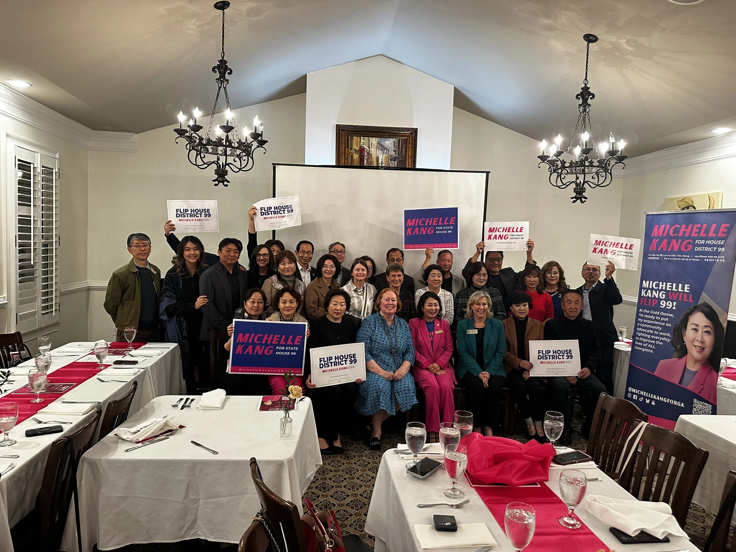 A group of people holding campaign signs for Michelle Kang, gathered in a banquet hall with tables set for dining, chandeliers hanging from the ceiling, and campaign posters on display.