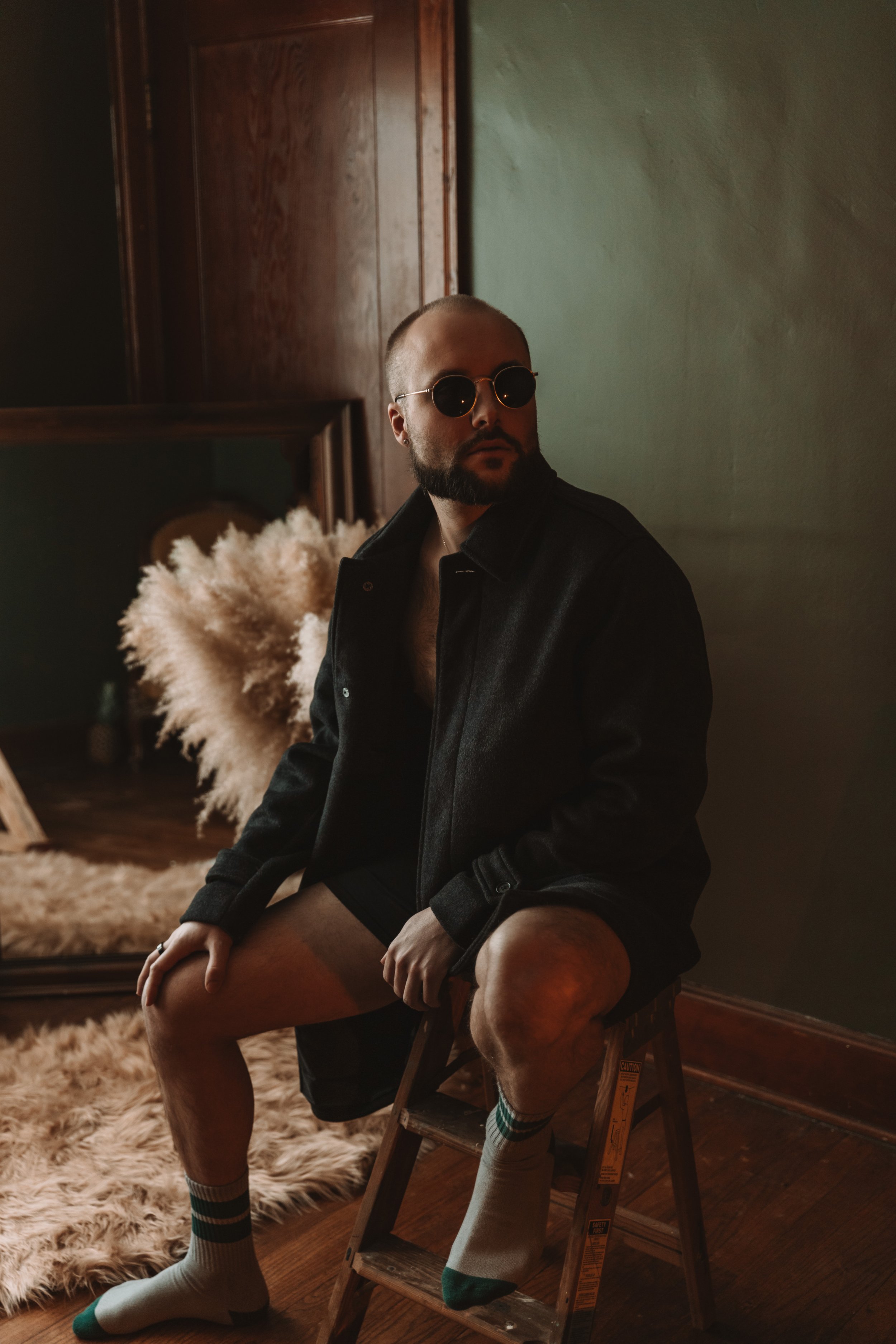 A man with sunglasses, beard, and shaved head sitting on a ladder, wearing a black jacket, shorts, and socks, indoors near a mirror and a furry rug.