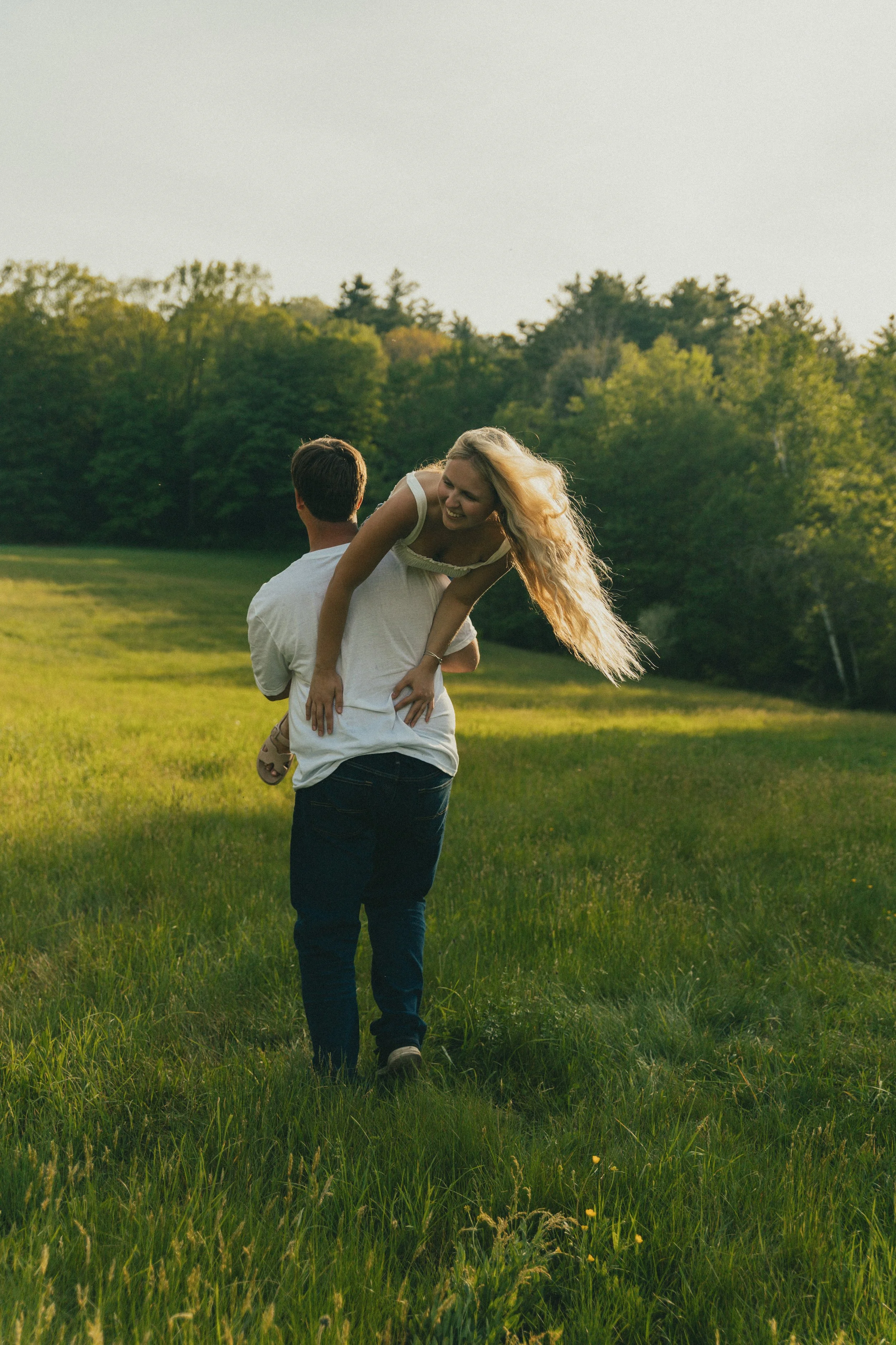 A man is carrying a woman on his back across a grassy field during sunset, with trees in the background.
