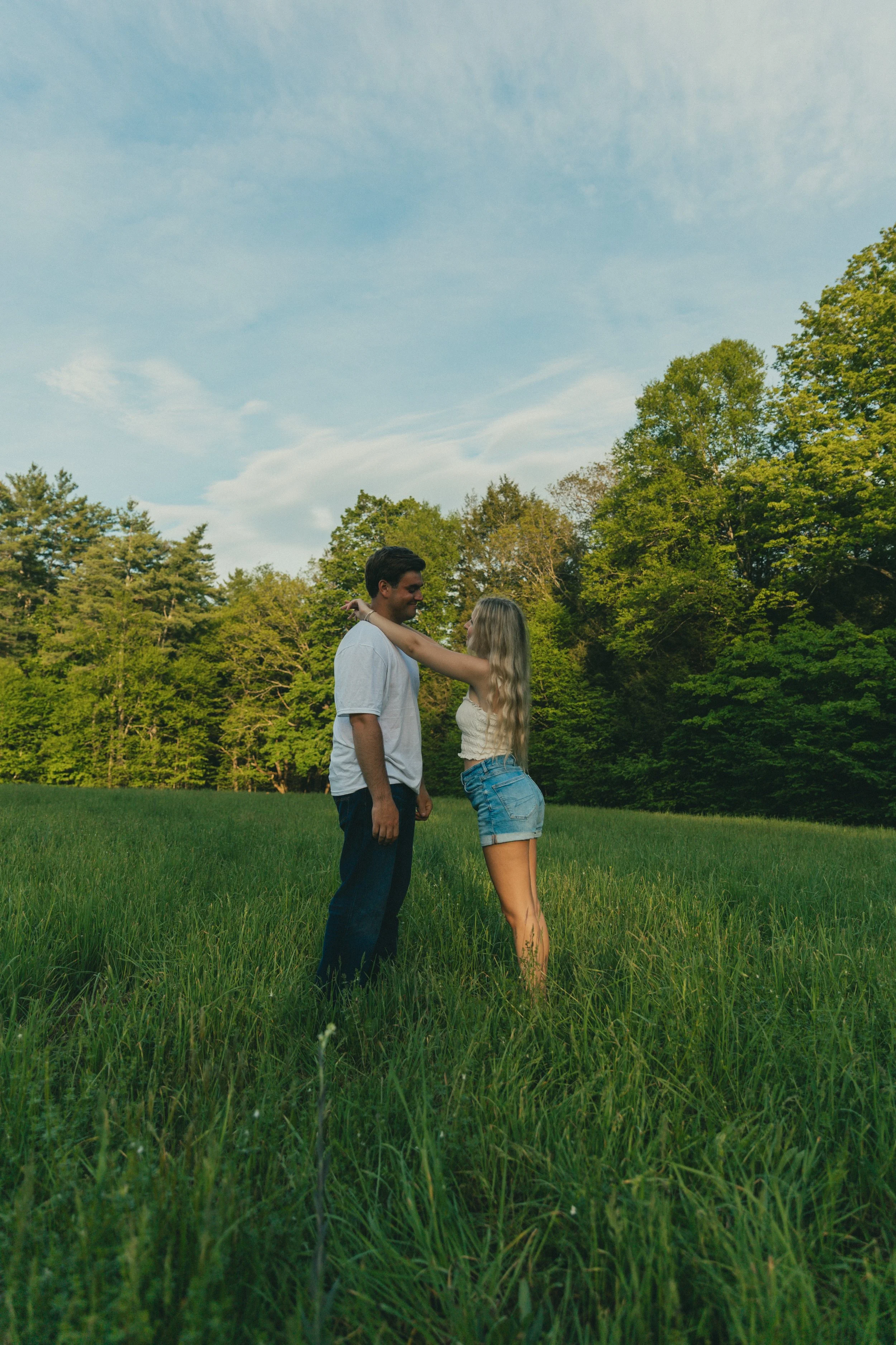 A young couple standing close together in a green grassy field, with trees and a blue sky with clouds in the background. Western Massachusetts