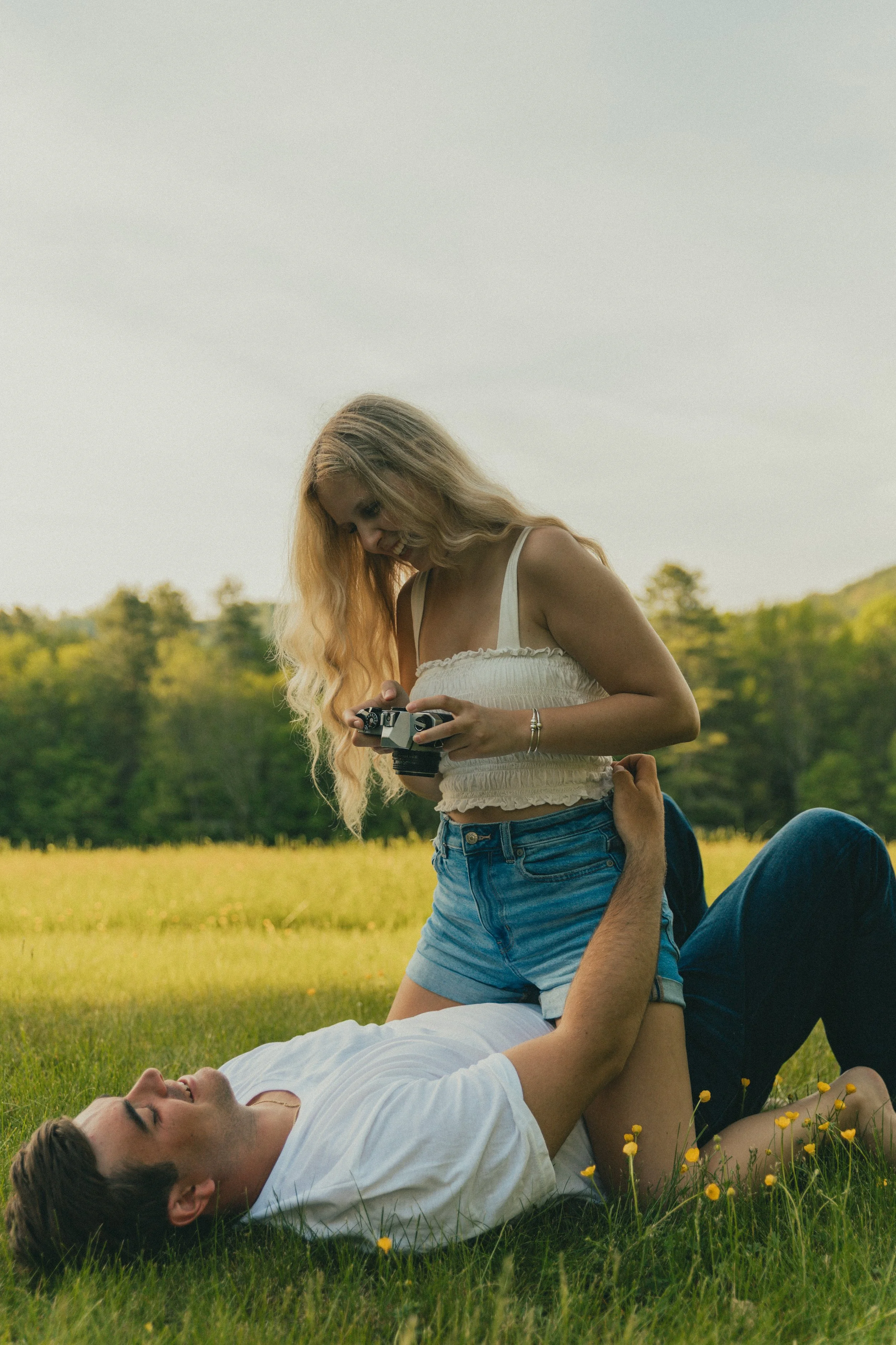 A young woman with long blonde hair standing on a grassy field, holding a camera, smiling and looking down. A young man lying on his back behind her, supporting her with his hands on her waist. They are outdoors with trees and a cloudy sky in the bac