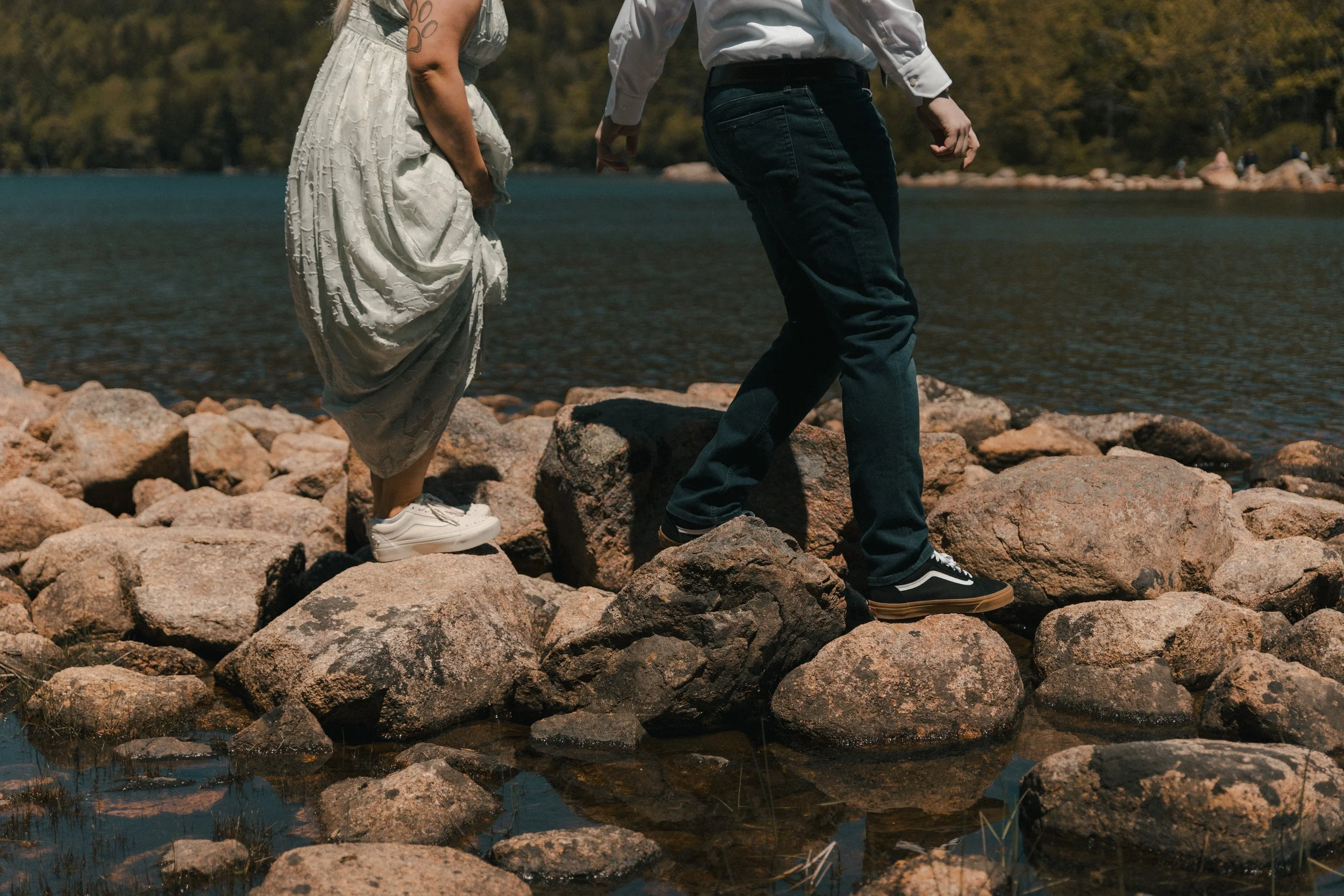 Close-up of a couple walking on rocks by a body of water, with mountains and trees in the background. elopement in Acadia National Park