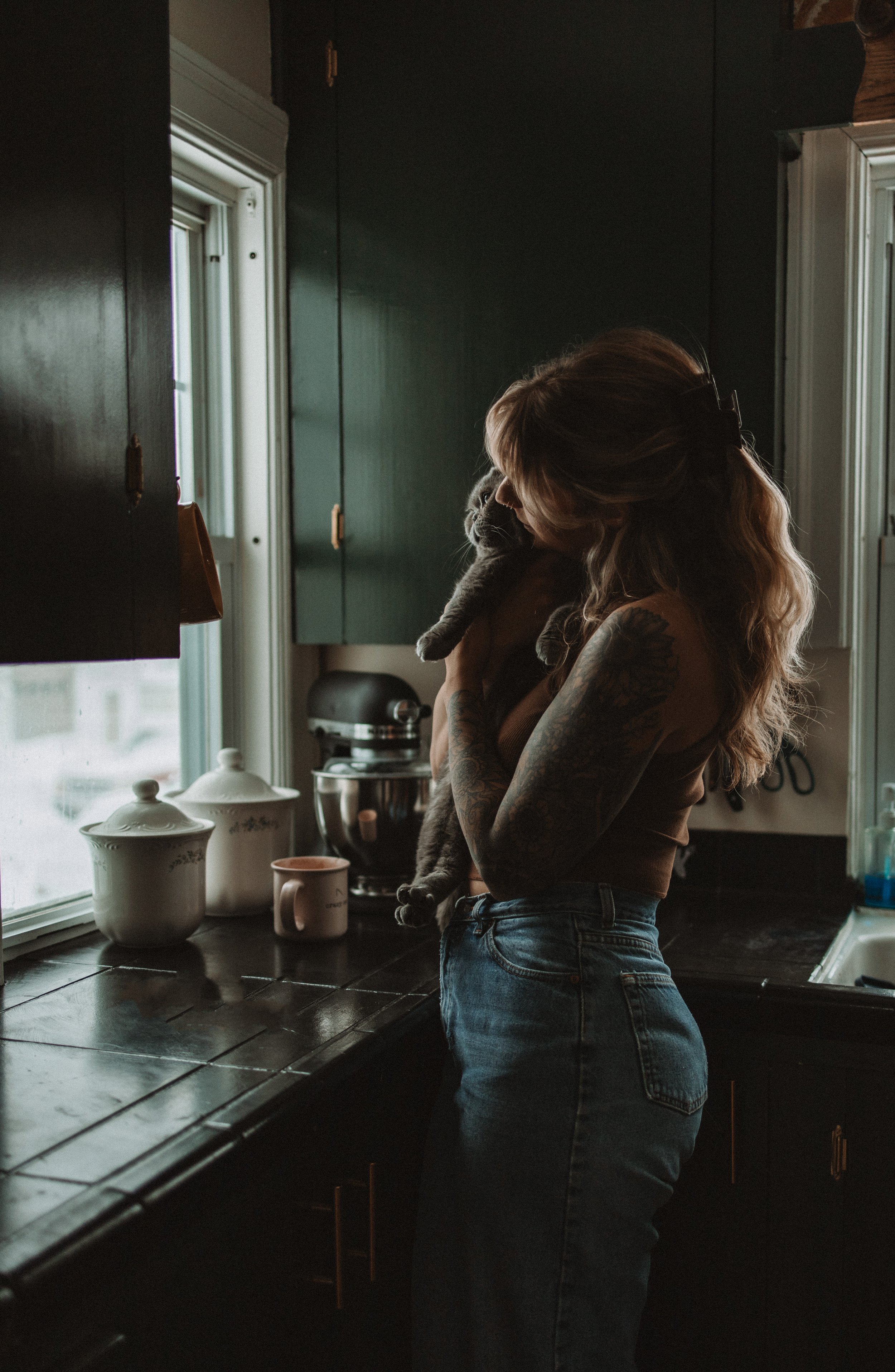 A woman with long, wavy hair and tattoos on her arms, holding a gray cat in a cozy kitchen near a window with natural light. Various kitchen items are on the counter, including a teapot, a mug, and a stand mixer.