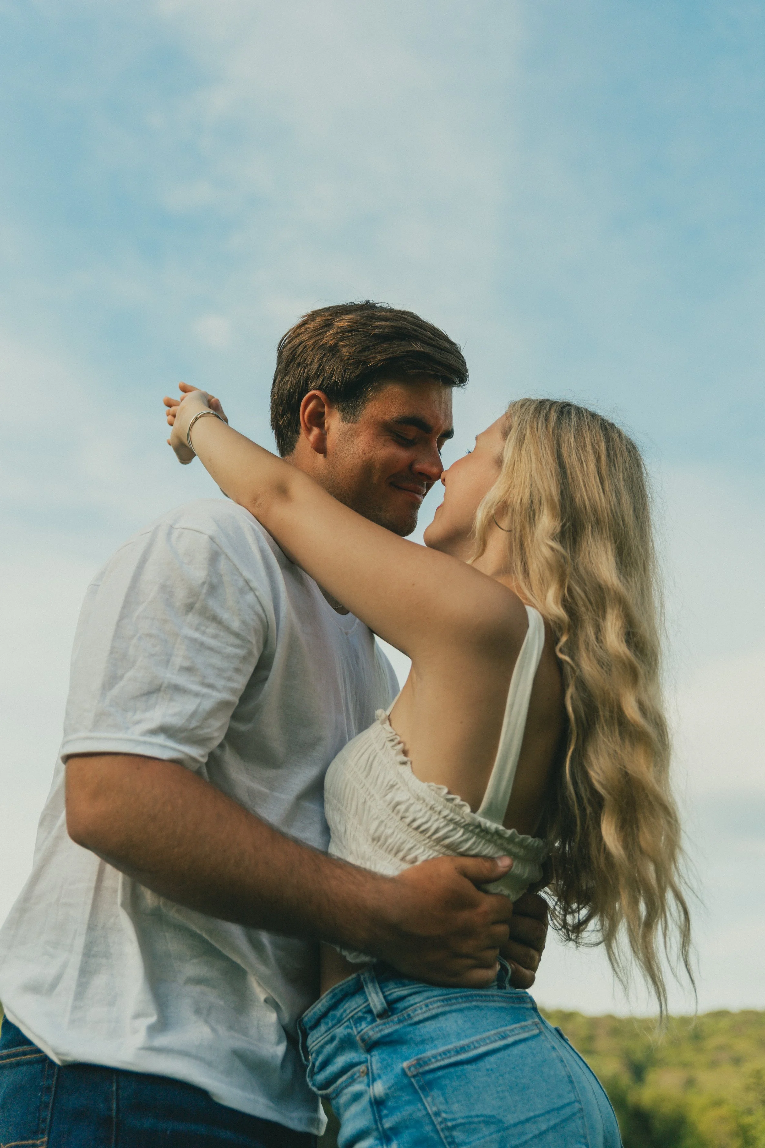 A man and woman embracing outdoors, standing close with foreheads touching, against a background of blue sky and green landscape.