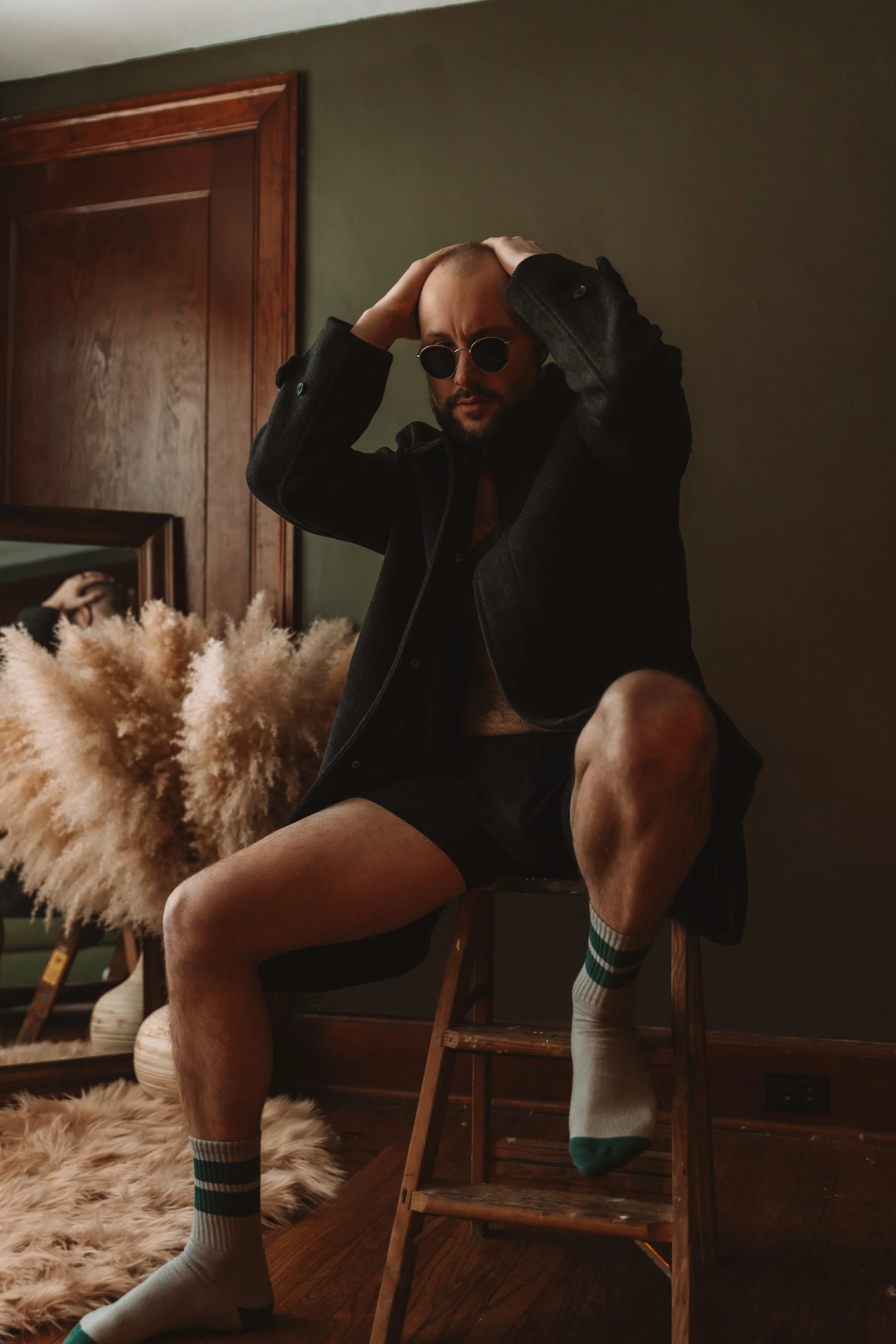 A man with a bald head and sunglasses sitting on a wooden step stool in a room, wearing a black coat, with one leg crossed over the other, wearing striped socks, and touching his head with both hands.
