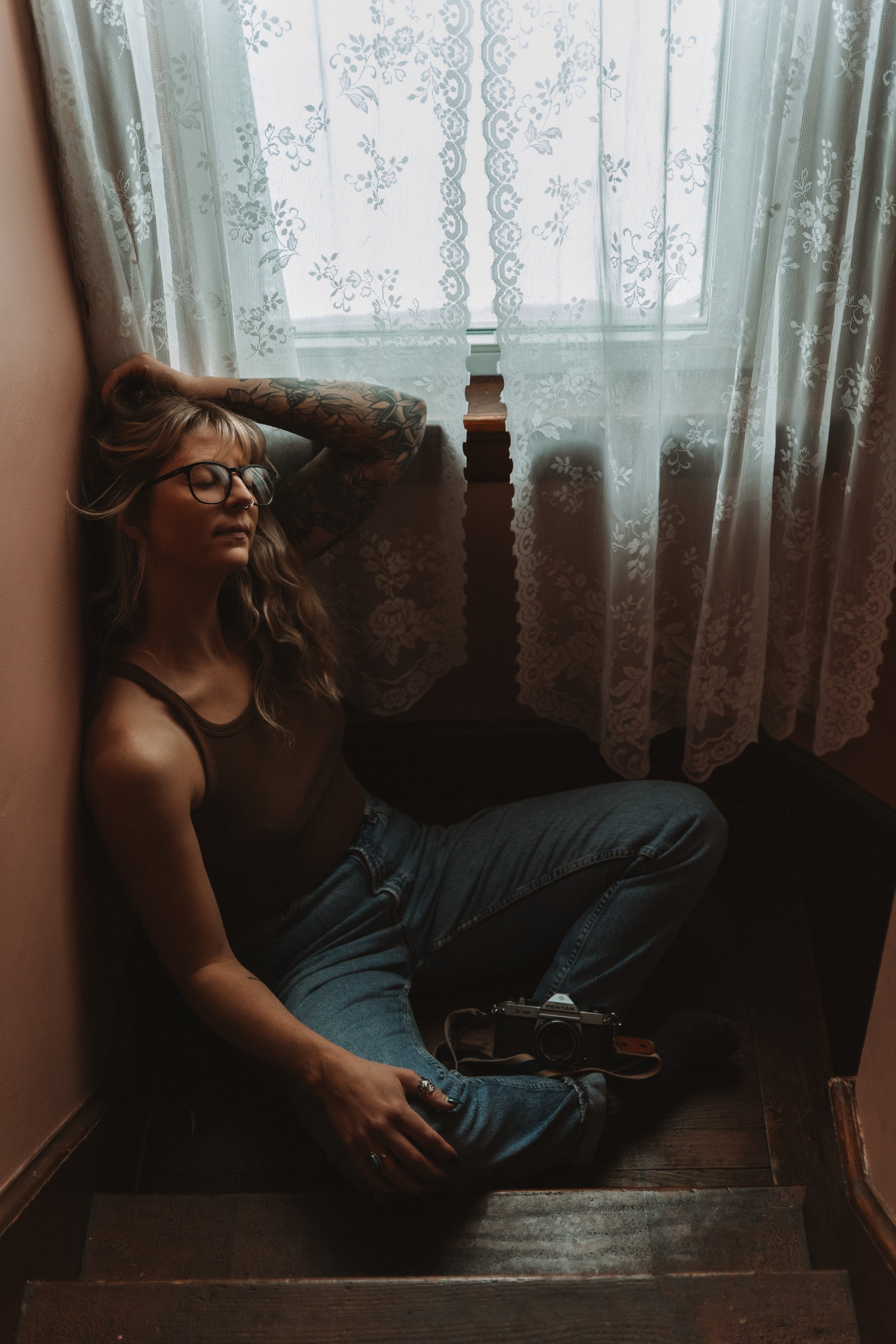 A young woman with glasses and tattoos is sitting on the floor by a staircase, leaning against the wall, with her eyes closed. She has her left arm raised above her head and is holding her hair, while her right arm rests on her knee. Sunlight filters through lace curtains on a window behind her, creating a soft, cozy atmosphere.