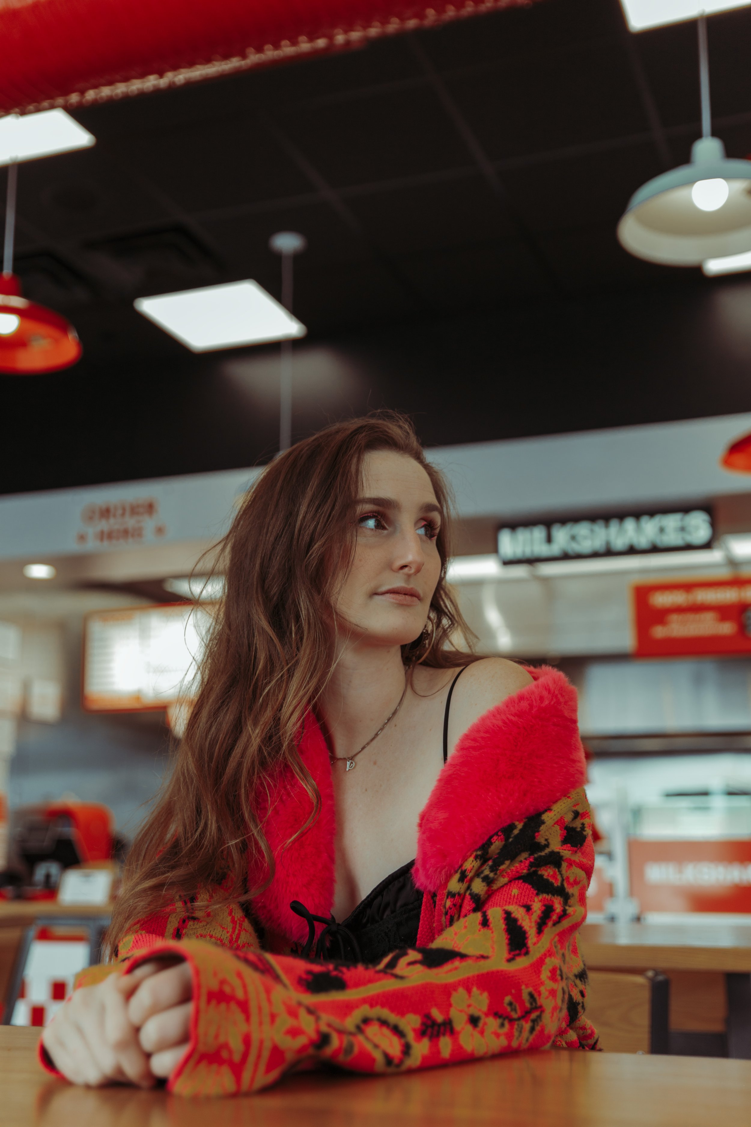 A young woman with long, wavy brown hair sitting at a table in a fast food restaurant, wearing a colorful jacket with red, black, and gold patterns and a black top with spaghetti straps. She is looking to her right with a slight smile.