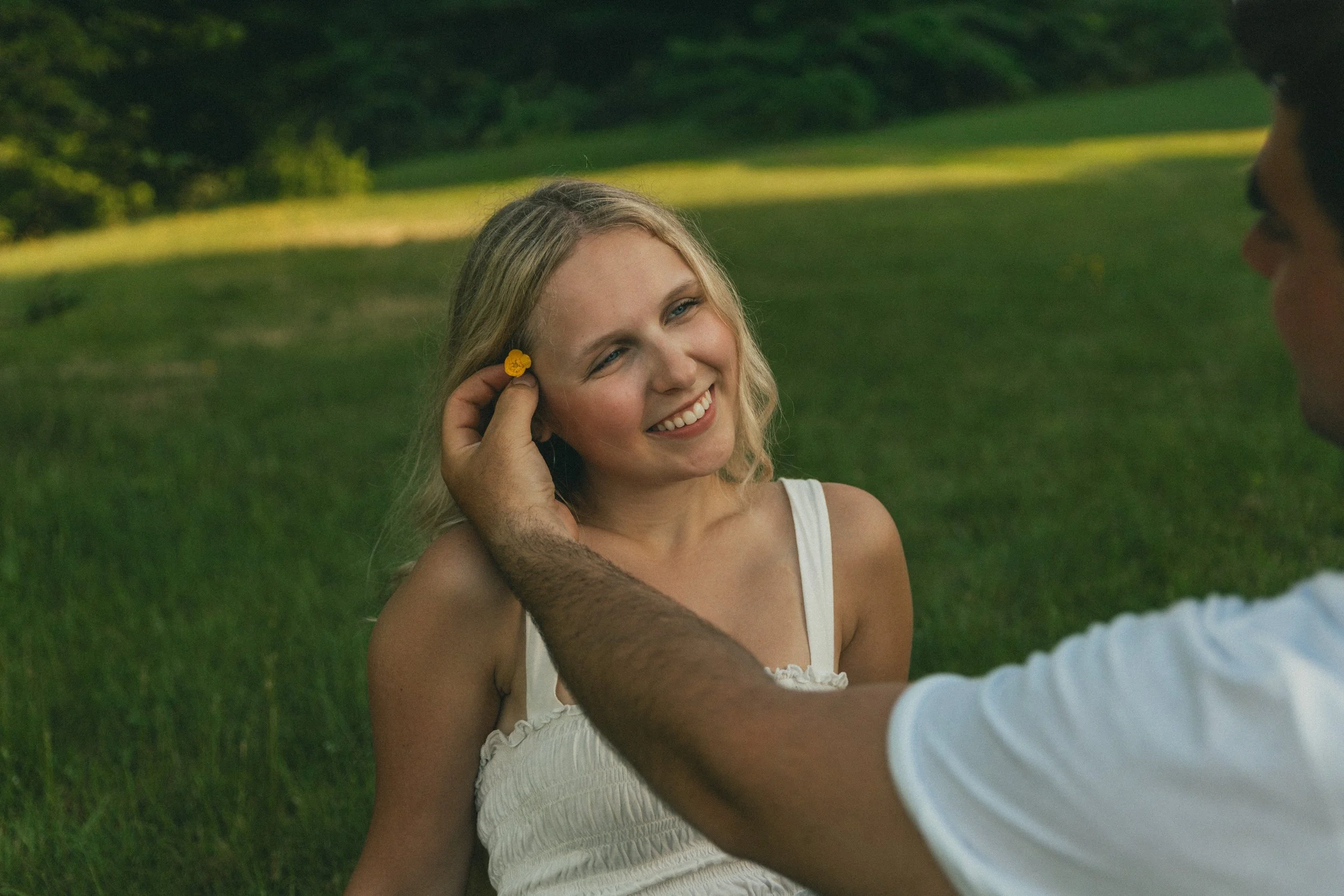 A woman with blonde hair in a white dress smiling as she is about to receive a yellow flower behind her ear from a person with darker skin, sitting outdoors on a grassy area with trees in the background.