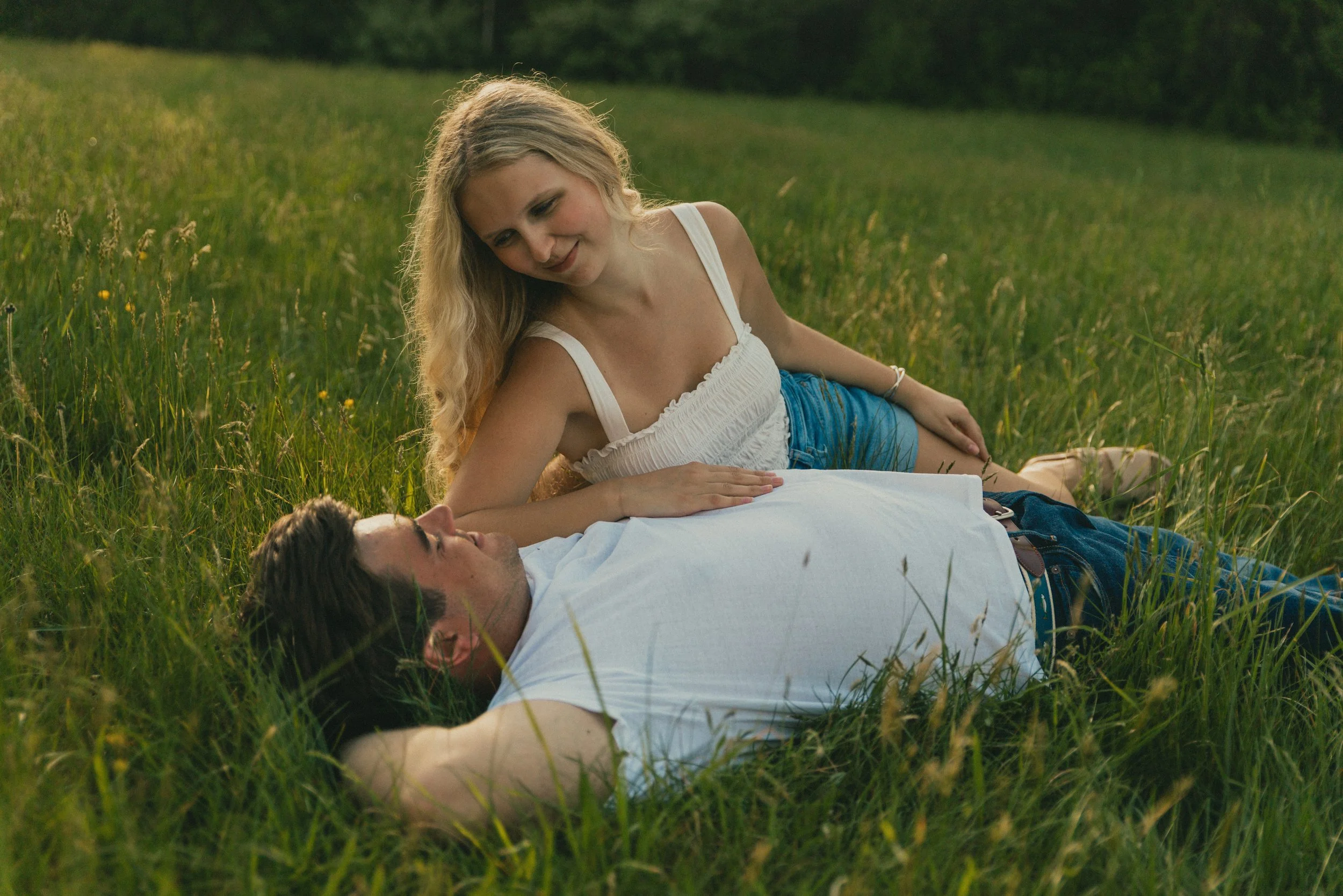 A woman with long blonde hair in a white sleeveless top sitting on the grass and smiling at a man lying down with his eyes closed, in a lush green field during the daytime.