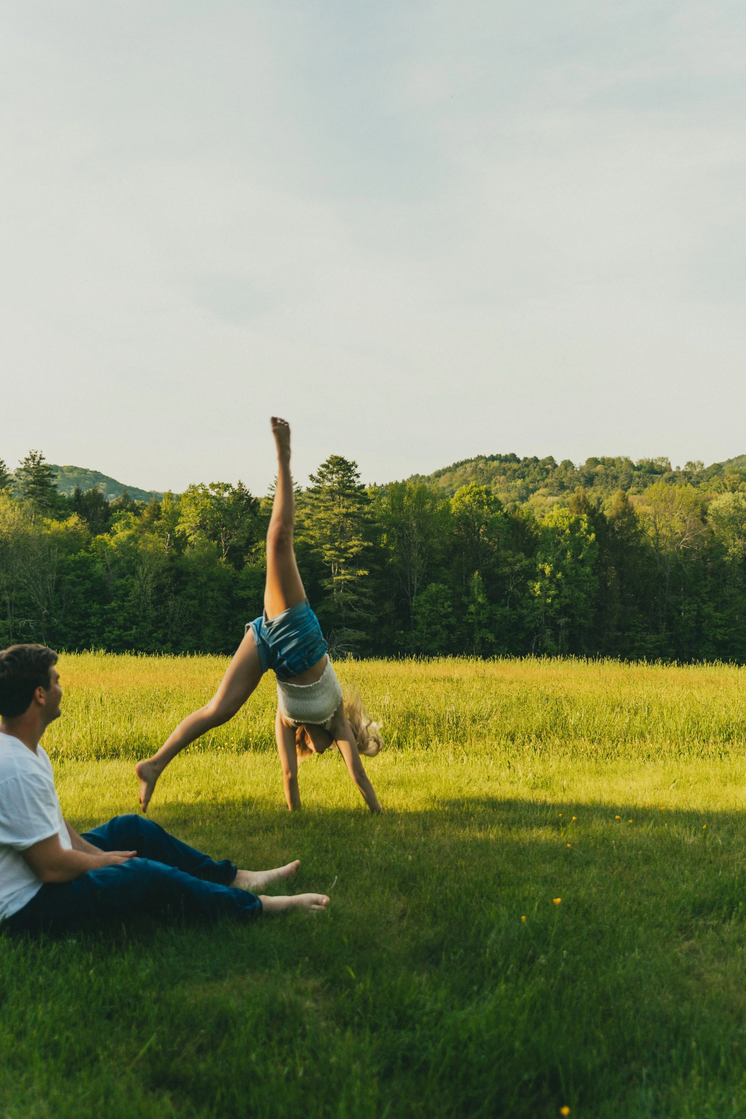 A girl doing a handstand on a grassy field while a boy in a white shirt and blue pants sits nearby, with a background of trees and hills under a clear sky.