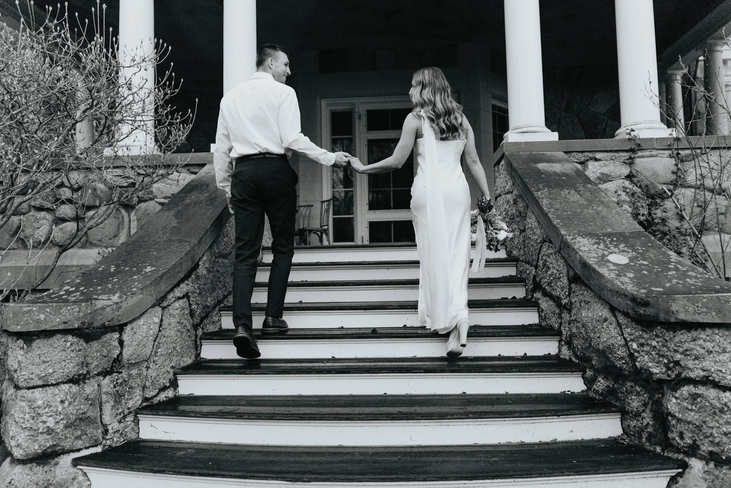 A black and white photo of a man and woman holding hands while walking up stairs outside a house. The man is dressed in a white shirt and dark pants, and the woman is wearing a white dress and carrying a bouquet of flowers.