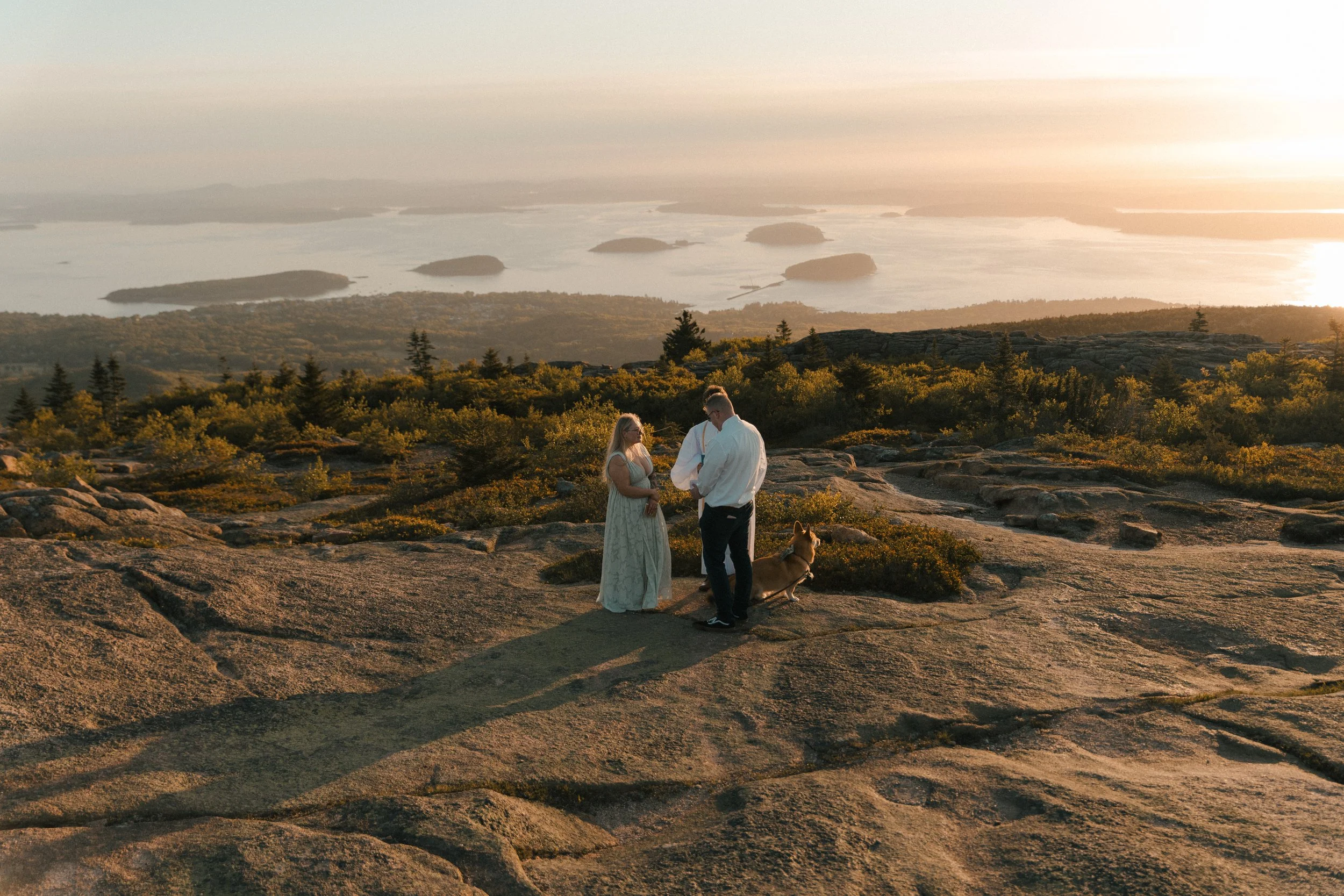 A couple dressed in white taking wedding vows on a rocky hilltop with a dog, overlooking a scenic landscape of lakes and islands at sunset.