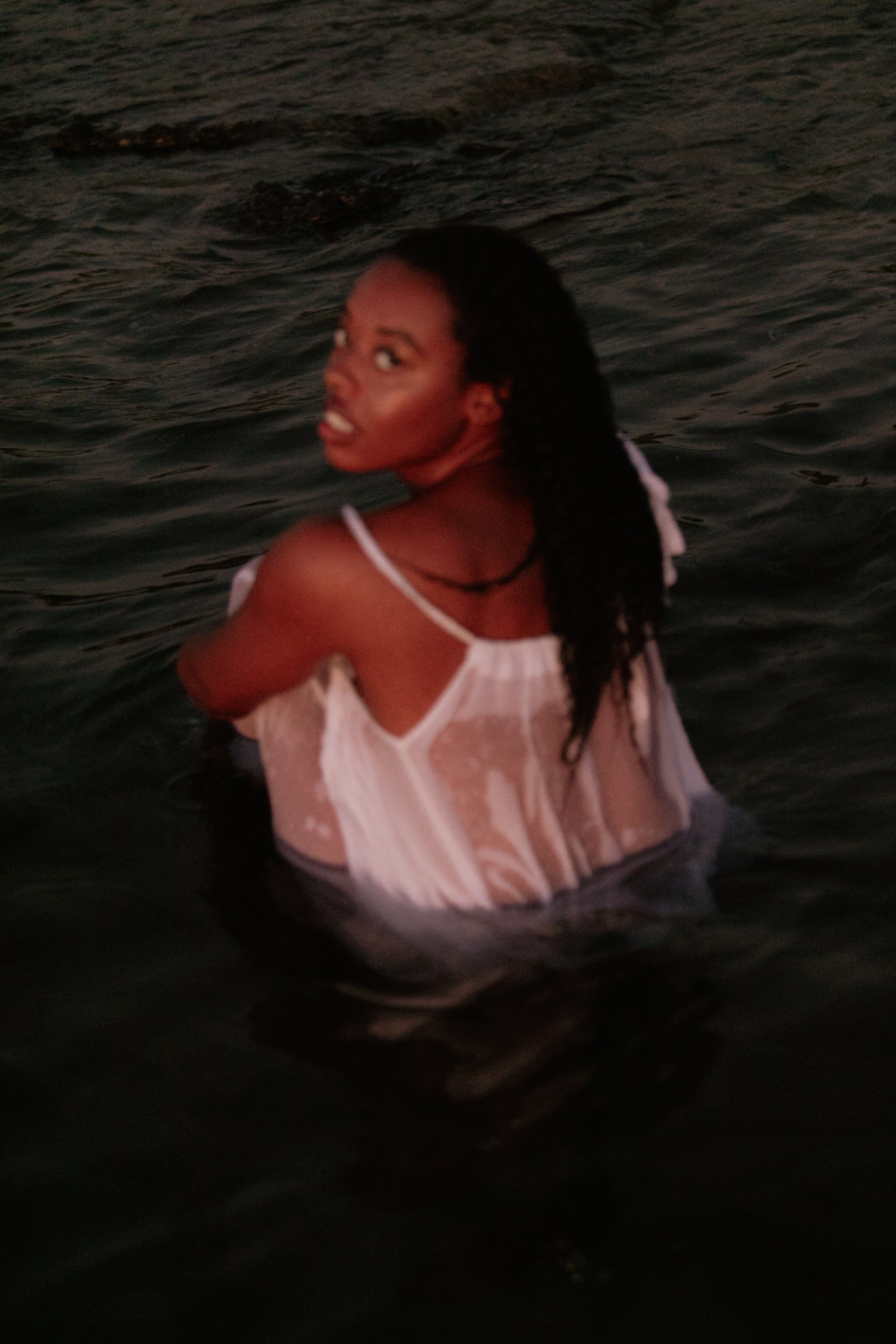 A woman with long, dark braided hair wearing a wet, white, semi-transparent dress is in the water at sunset, looking back over her shoulder.