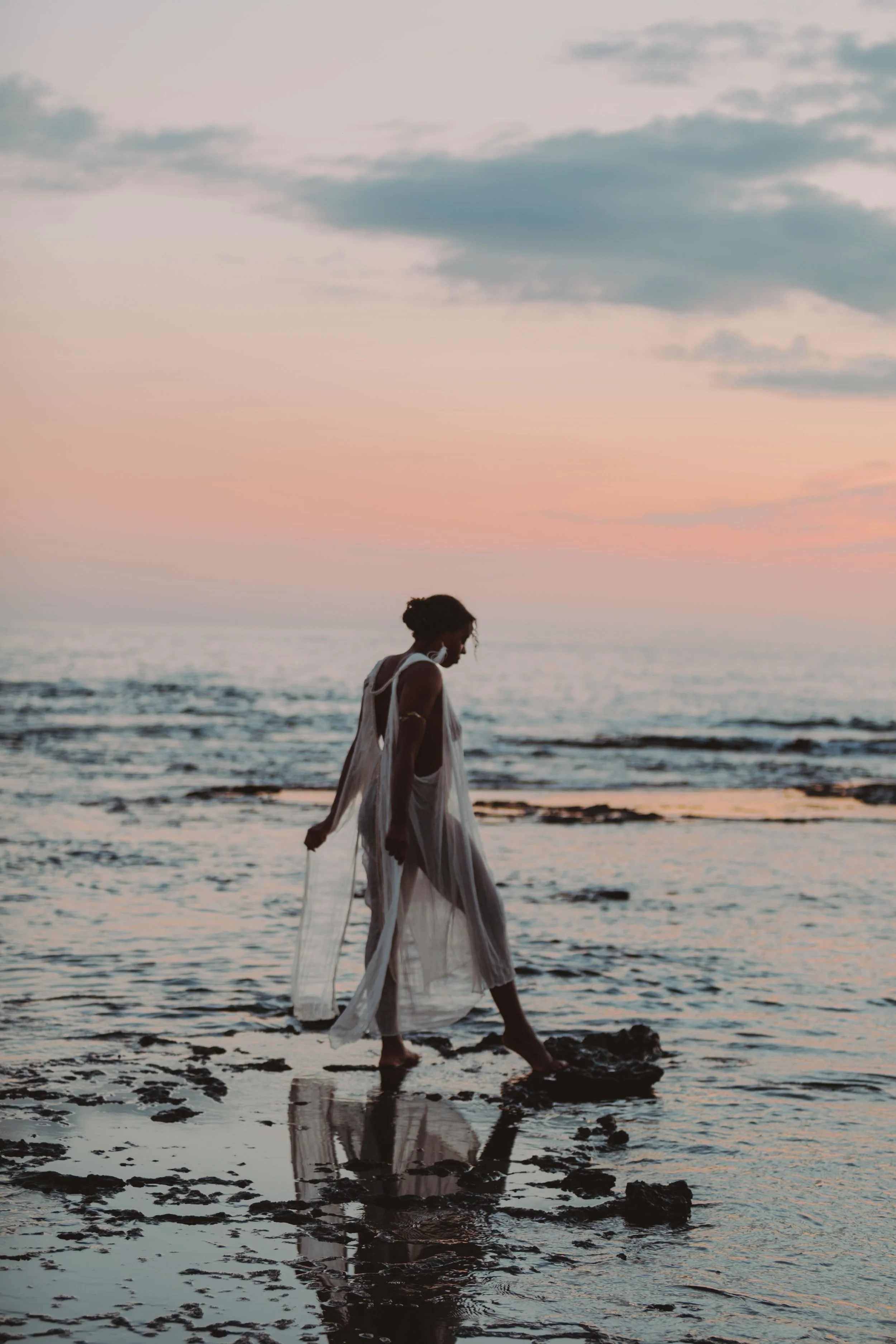A woman walking in shallow water at the beach during sunset, wearing a flowing white dress.