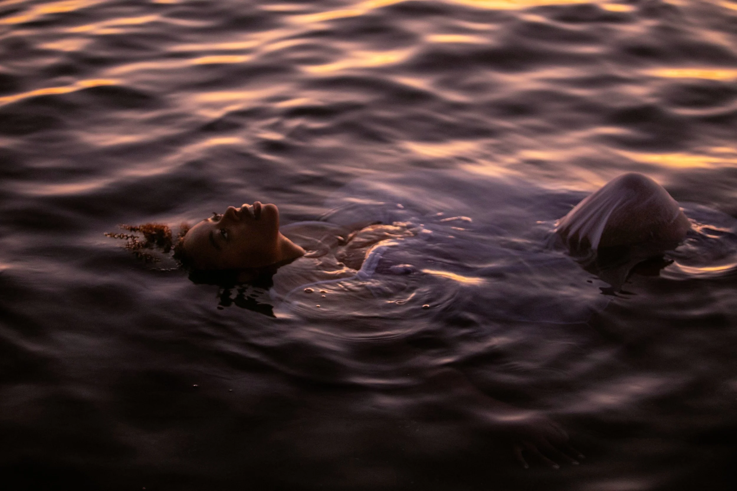 A woman floating on her back in water at sunset, her face and upper body visible with a peaceful expression.
