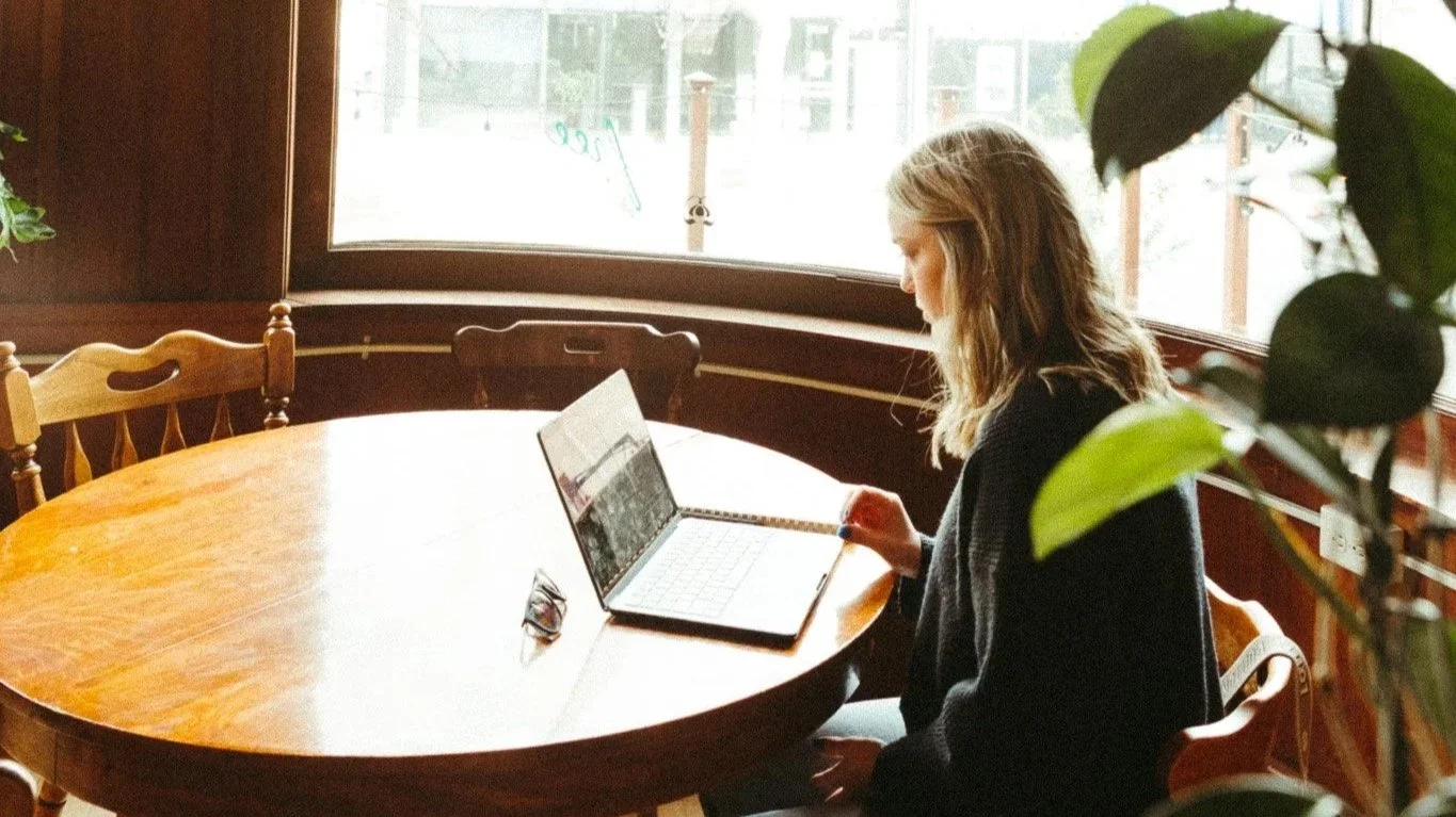 A woman with shoulder-length blonde hair sitting at a round wooden table in a cozy café, working on a laptop with a pair of glasses nearby. The café has large windows, letting in natural light, and green plants are partially visible in the foreground.