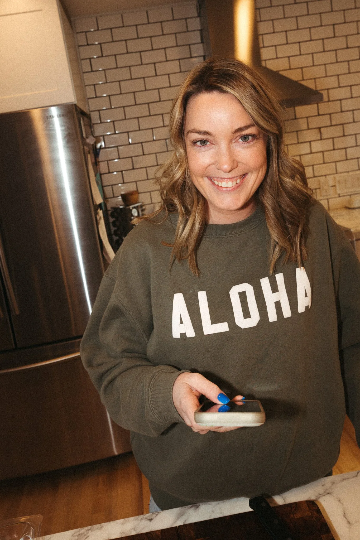 A woman with wavy brown hair wearing a green sweatshirt that says 'ALOHA' stands in a kitchen, smiling at the camera while holding a smartphone.