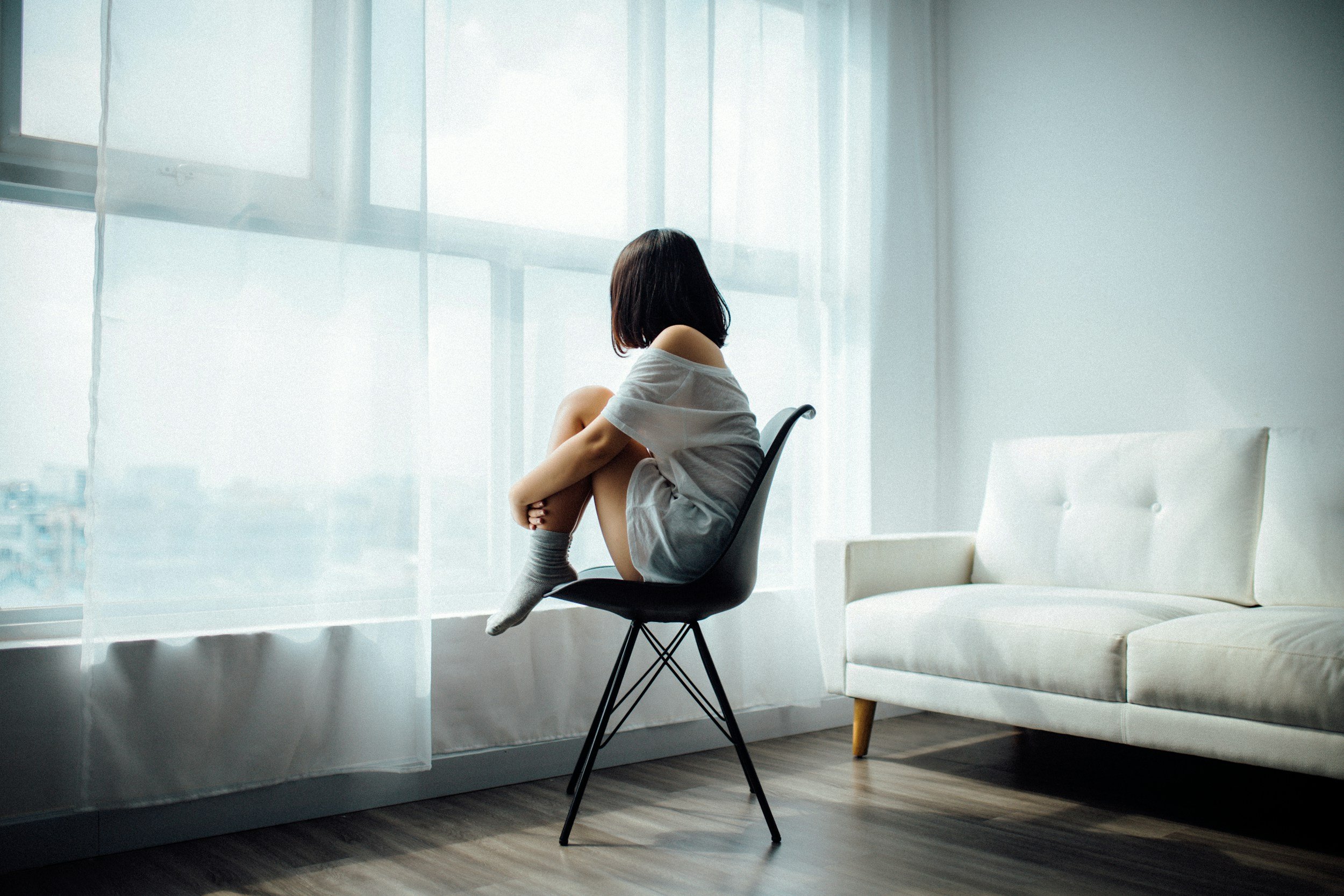 A woman with short dark hair sitting alone on a black chair in a bright room with large windows and sheer curtains, looking out. Who may benefit from a from therapy provided by Empowering Hope and our skilled therapists.