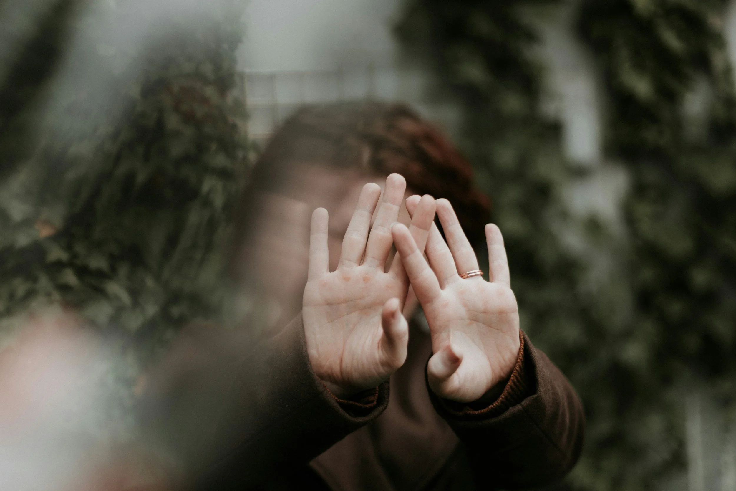 Two hands pressing against a glass surface, with a blurred background and a person with reddish-brown hair behind the glass.  An Empowering Hope therapist could support her with her mental health needs.