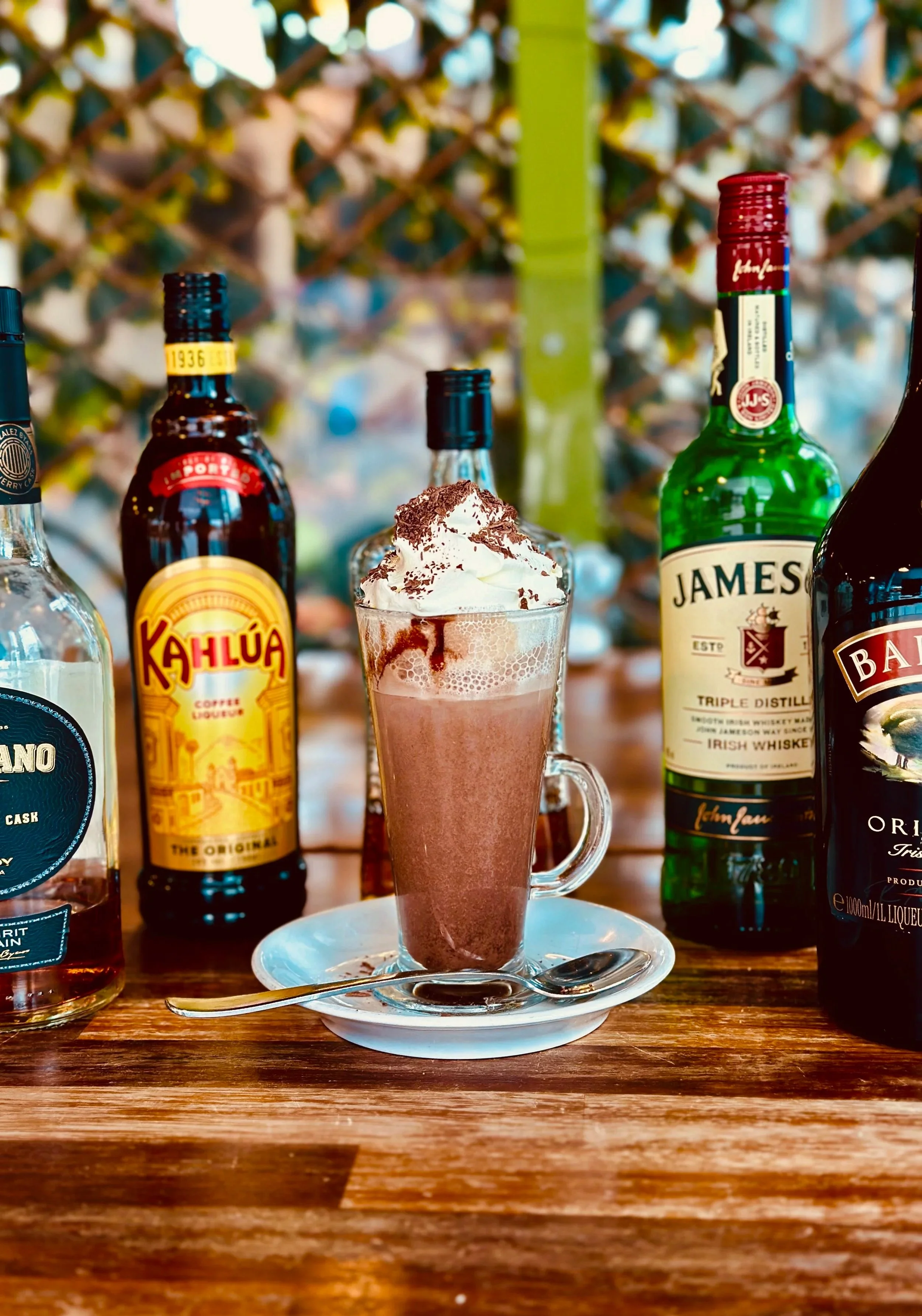 A glass of whipped cream-topped chocolate drink on a saucer with a spoon, surrounded by bottles of Jameson Irish whiskey, Kahluá coffee liqueur, and other bottles on a wooden table.