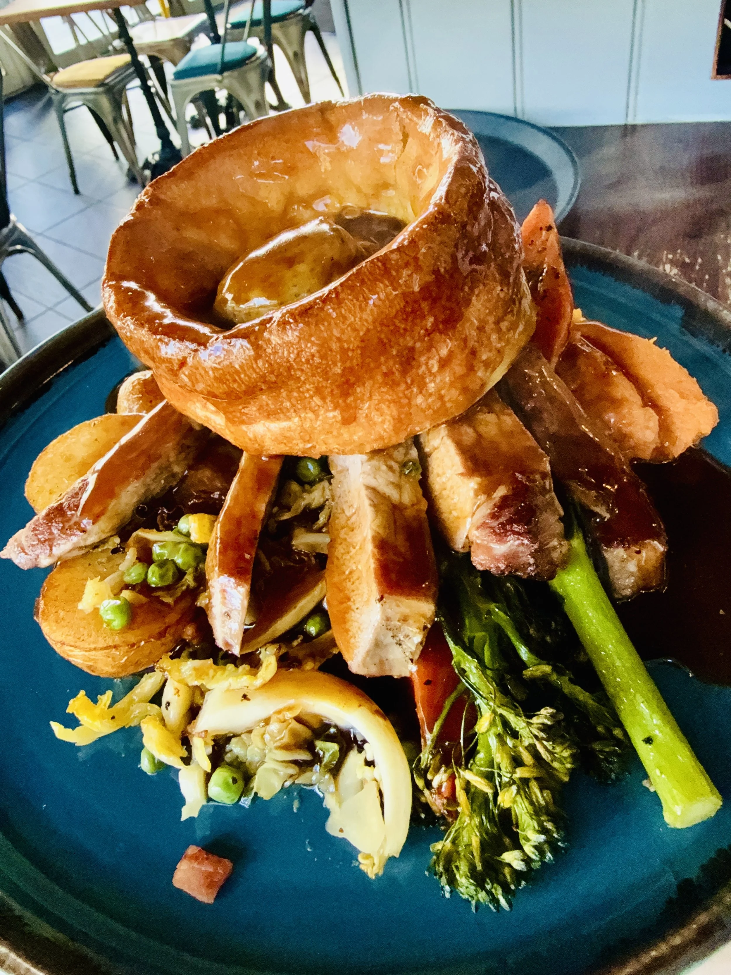 A plated dish featuring a savory bread bowl, sliced roasted meats, vegetables including broccoli, carrots, peas, and artichoke, all topped with gravy, served on a dark plate in a restaurant setting.