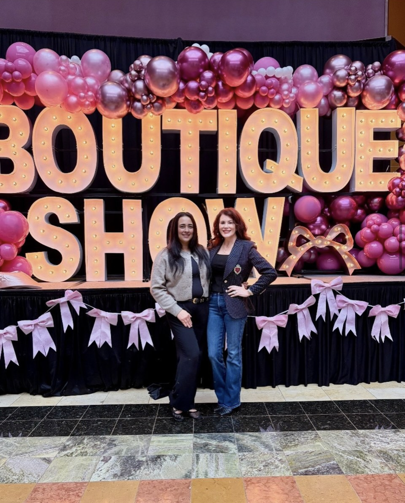 Two women standing in front of a decorative display with large illuminated letters spelling "BOUTIQUE SHOW" surrounded by pink and purple balloons, with pink bows and ribbon garlands.