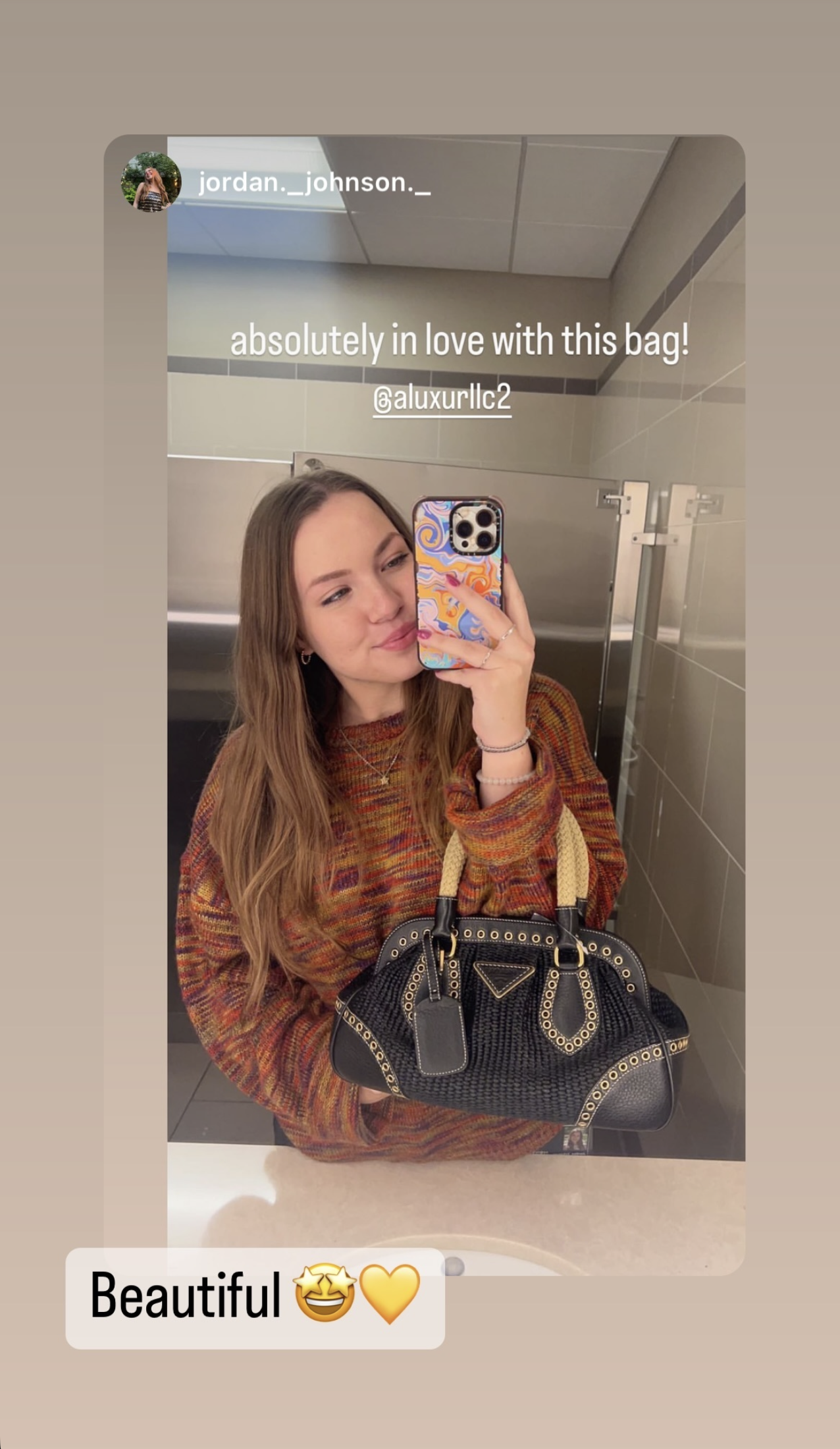 A young woman with long light brown hair taking a selfie in a bathroom mirror, holding a black designer handbag. She is wearing a multicolored plaid shirt and smiling softly. The background shows gray tiled walls and part of a metal bathroom stall. Text overlay on the image says, 'absolutely in love with this bag! @aluxurllc2' and a caption below says, 'Beautiful 😅💛'.