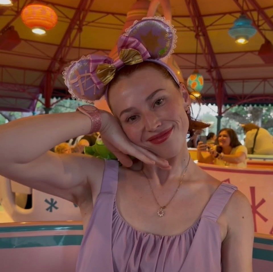 A woman with red hair smiling and resting her face on her hand. She is wearing a lavender dress, a necklace, and a headband with a large purple bow. The background shows a colorful carousel with hanging lanterns and a woman taking a photo.