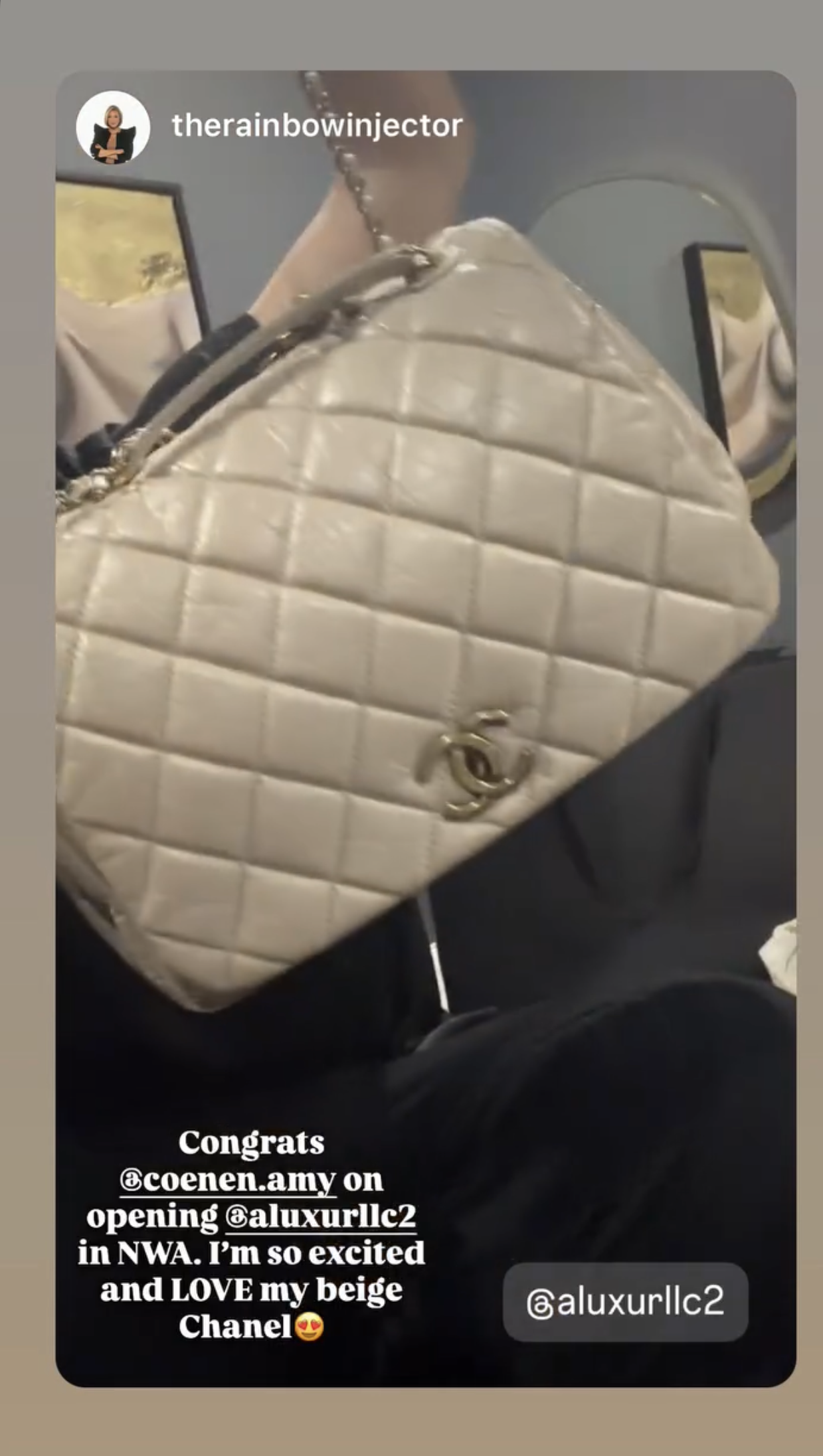 Close-up of a beige Chanel purse with quilted design, gold chain strap, and gold logo, held by a person. There are picture frames on the wall in the background.