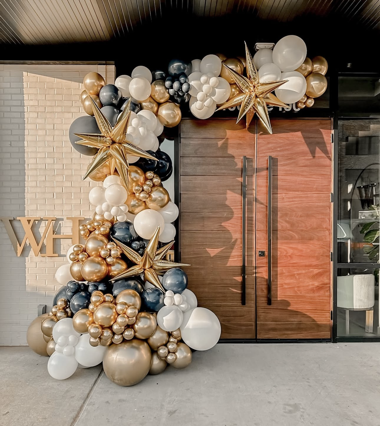 Decorative balloon arch with gold, white, black, and metallic balloons surrounding a wooden door, with a partial 'W' visible on the brick wall nearby.