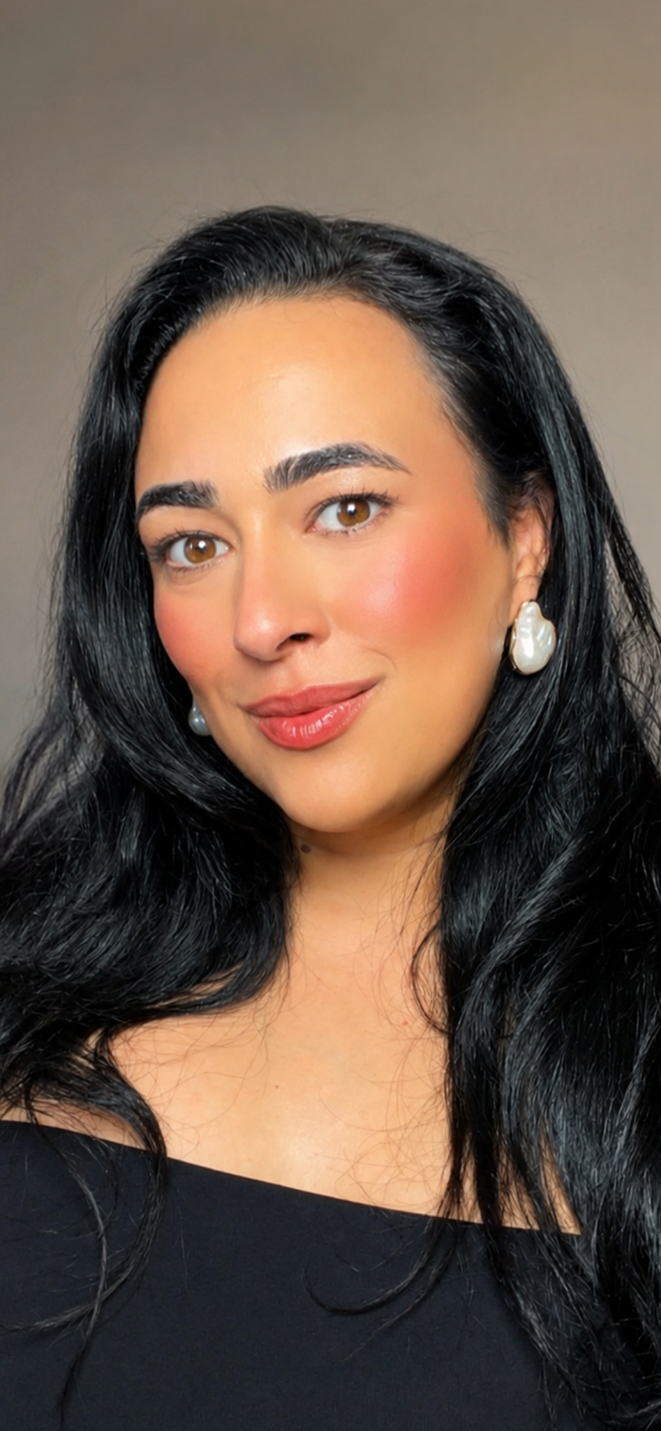 Close-up portrait of a woman with long black hair, brown eyes, and wearing pearl earrings, smiling softly.