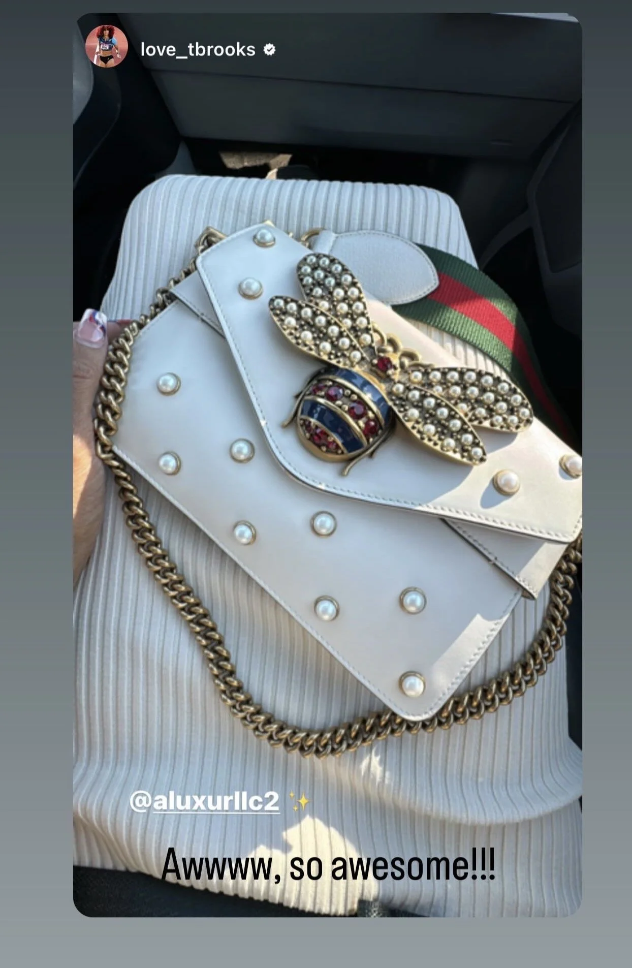 A white handbag with large pearls and a gold chain strap, placed on a light-colored striped garment in a car. A decorated bee brooch with pearls and rhinestones is attached to the bag, and a green and red striped strap is also visible.