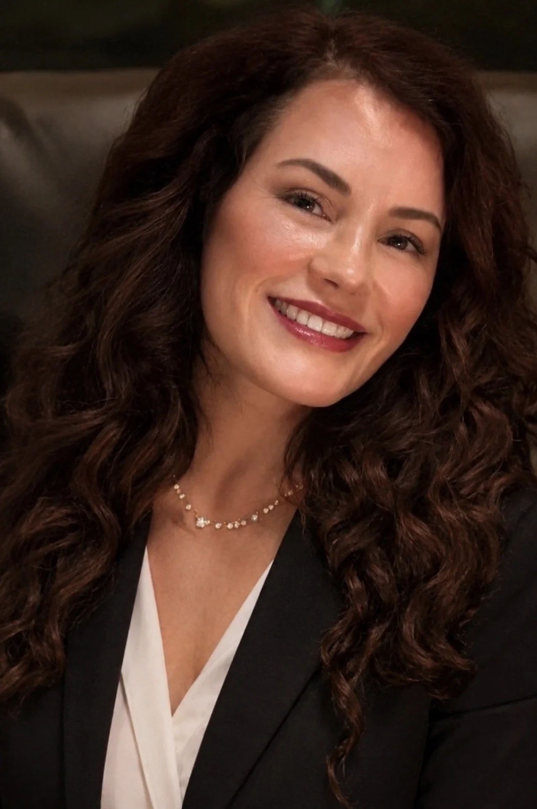 A woman with long curly brown hair, smiling and wearing a black blazer, white blouse, and a delicate necklace, seated in a dimly lit room.