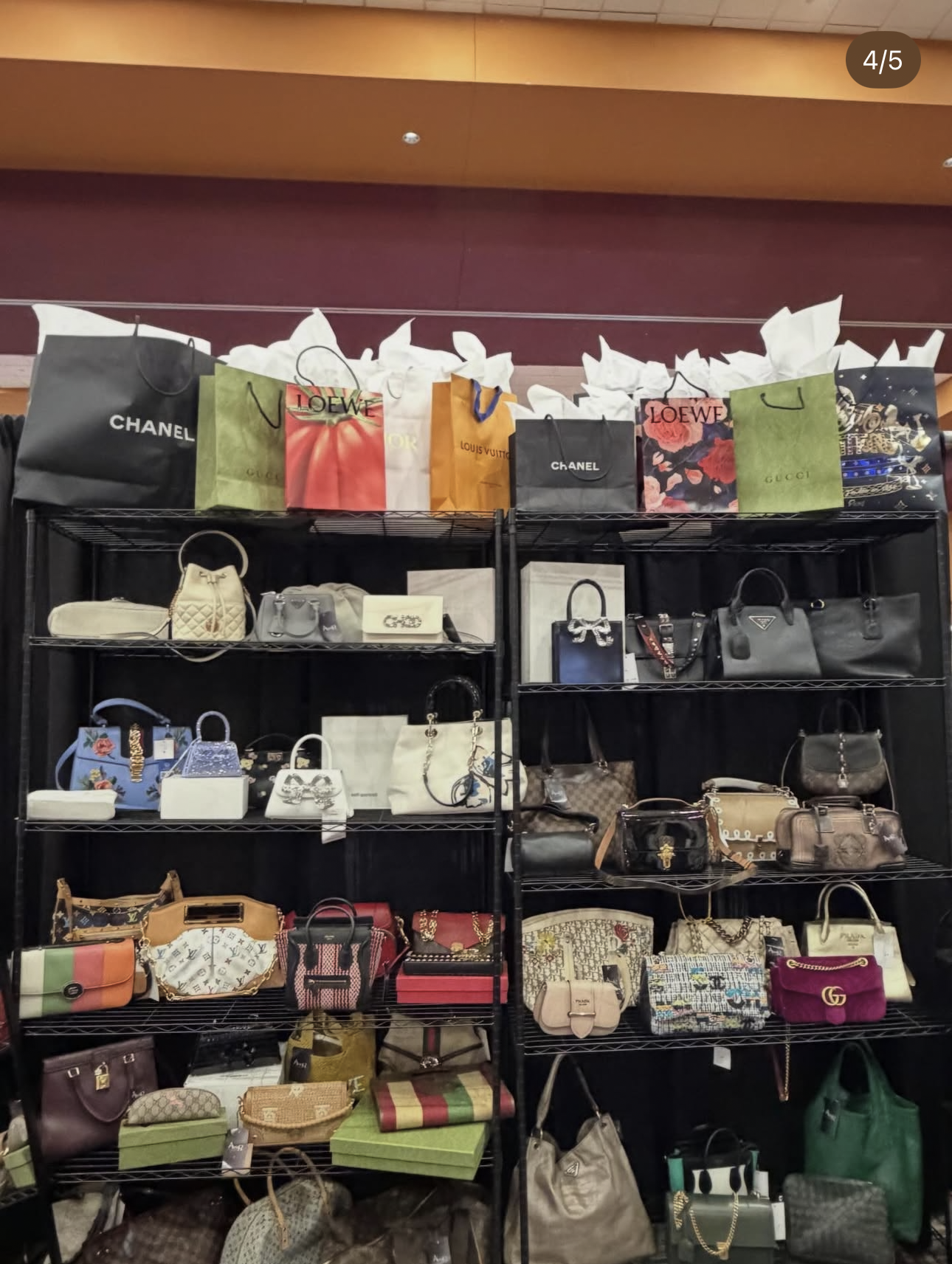 Shop display with multiple luxury handbags and shopping bags on black metal shelves. The top shelf has shopping bags from brands like Chanel, Loewe, Louis Vuitton, and Gucci. The lower shelves contain various designer handbags in different styles and colors.