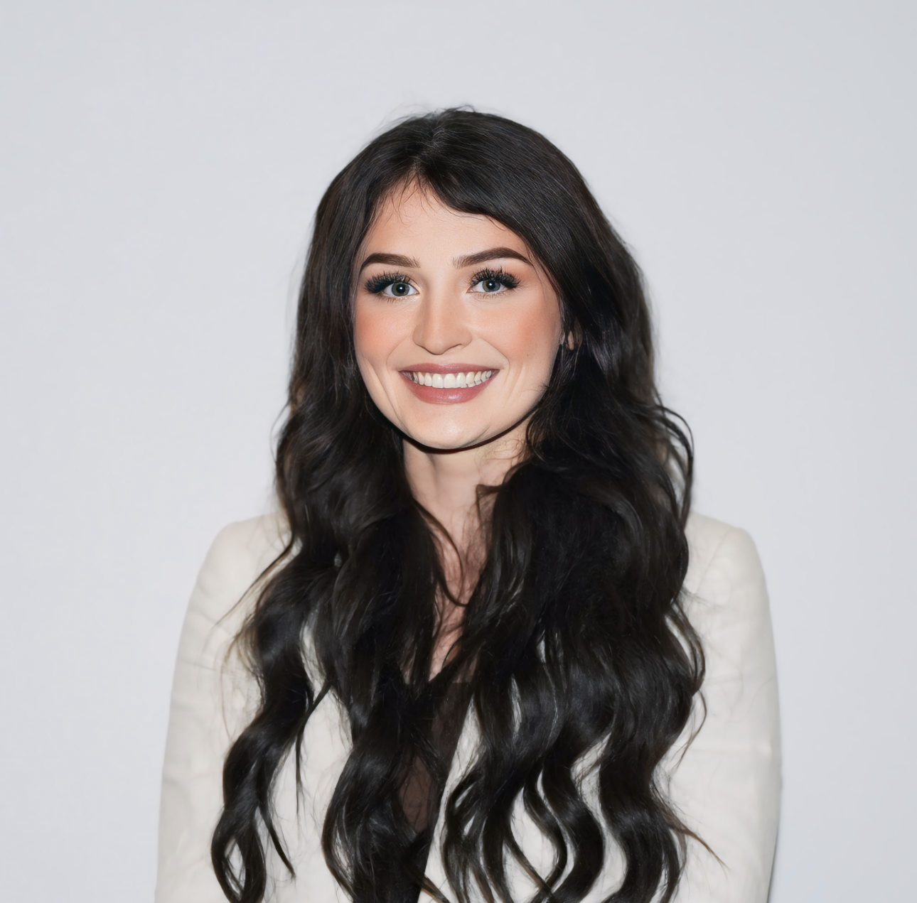 Portrait of a young woman with long, wavy dark hair smiling at the camera, wearing a white blazer against a plain light background.