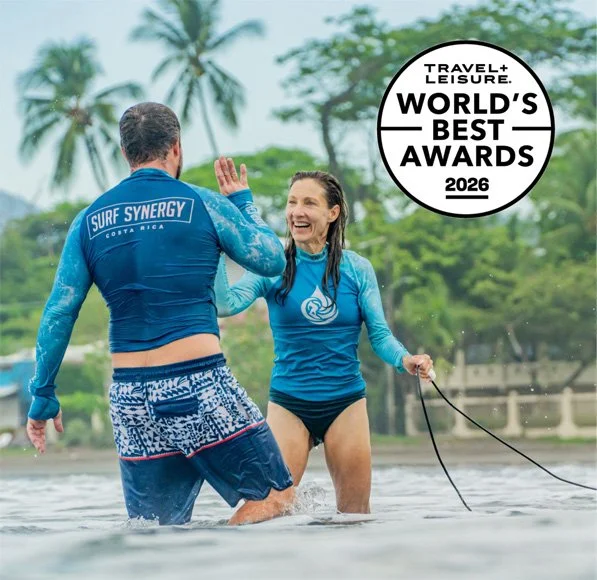 Two people in blue wetsuits high-fiving in a tropical beach setting, with a woman holding a leash attached to a dog. A circular logo in the top right corner reads 'Travel + Leisure World's Best Awards 2026'.