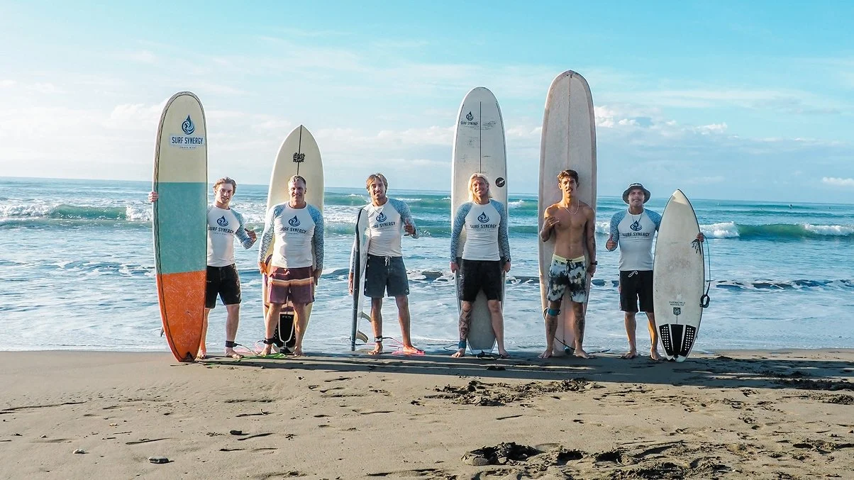 Group of six surfers standing on a Costa Rican beach with surfboards, wearing wetsuits or board shorts, near the ocean with waves and blue sky.