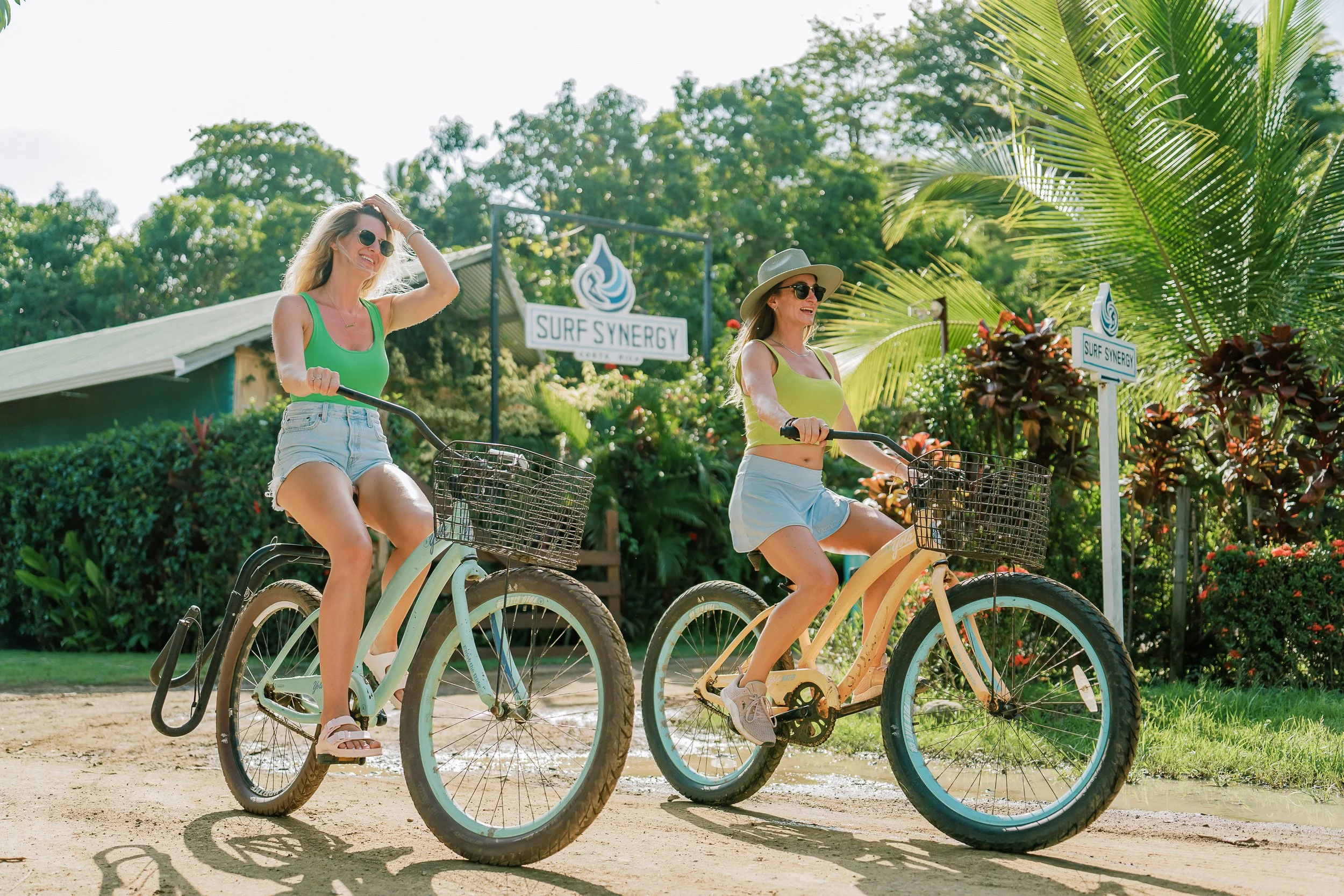 Two women riding bicycles on a dirt path, surrounded by lush greenery and tropical plants, with a sign that reads 'Surf Synergy' in the background.